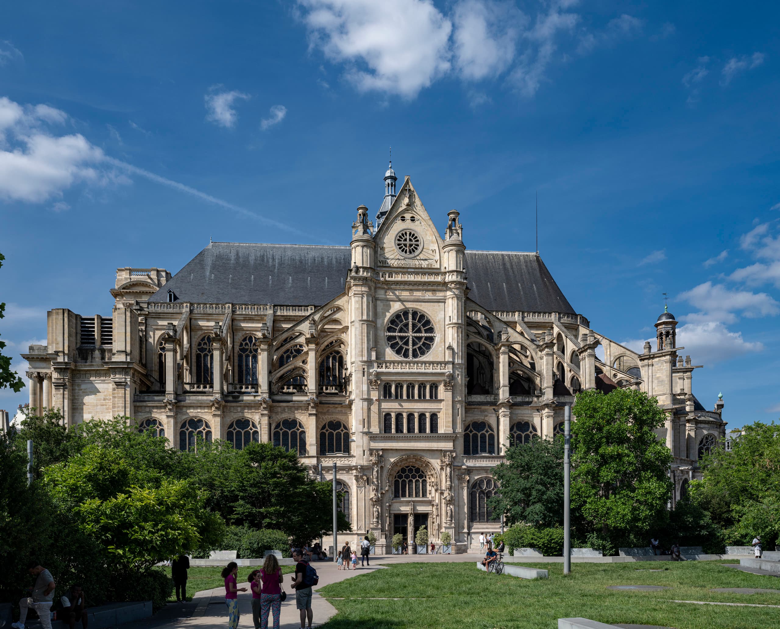 Außenansicht einer großen historischen Kirche mit aufwendiger Steinarchitektur, darunter Bogenfenster, Strebepfeiler und eine zentrale Rosette. Das Gebäude erhebt sich vor einem blauen Himmel mit vereinzelten Wolken und ist von grünen Bäumen sowie einem öffentlichen Park umgeben, in dem mehrere Menschen auf den Wegen spazieren gehen.