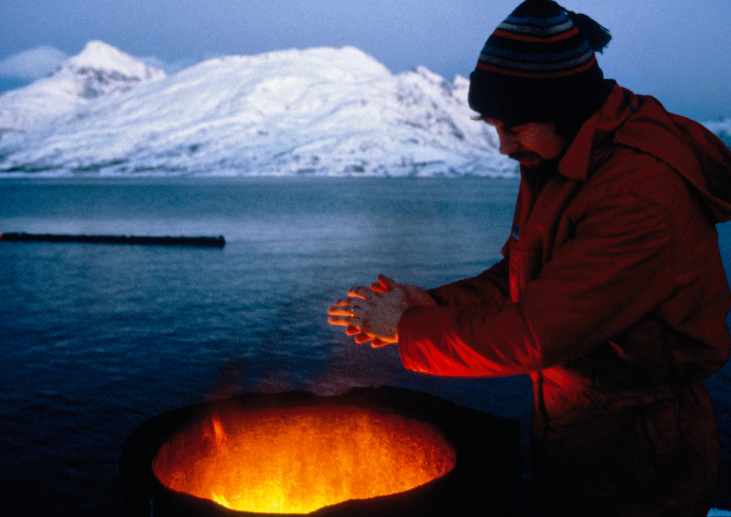 Mann wärmt sich am Feuer vor verschneiter Bergkulisse, Lichtdesign-Konzept.
