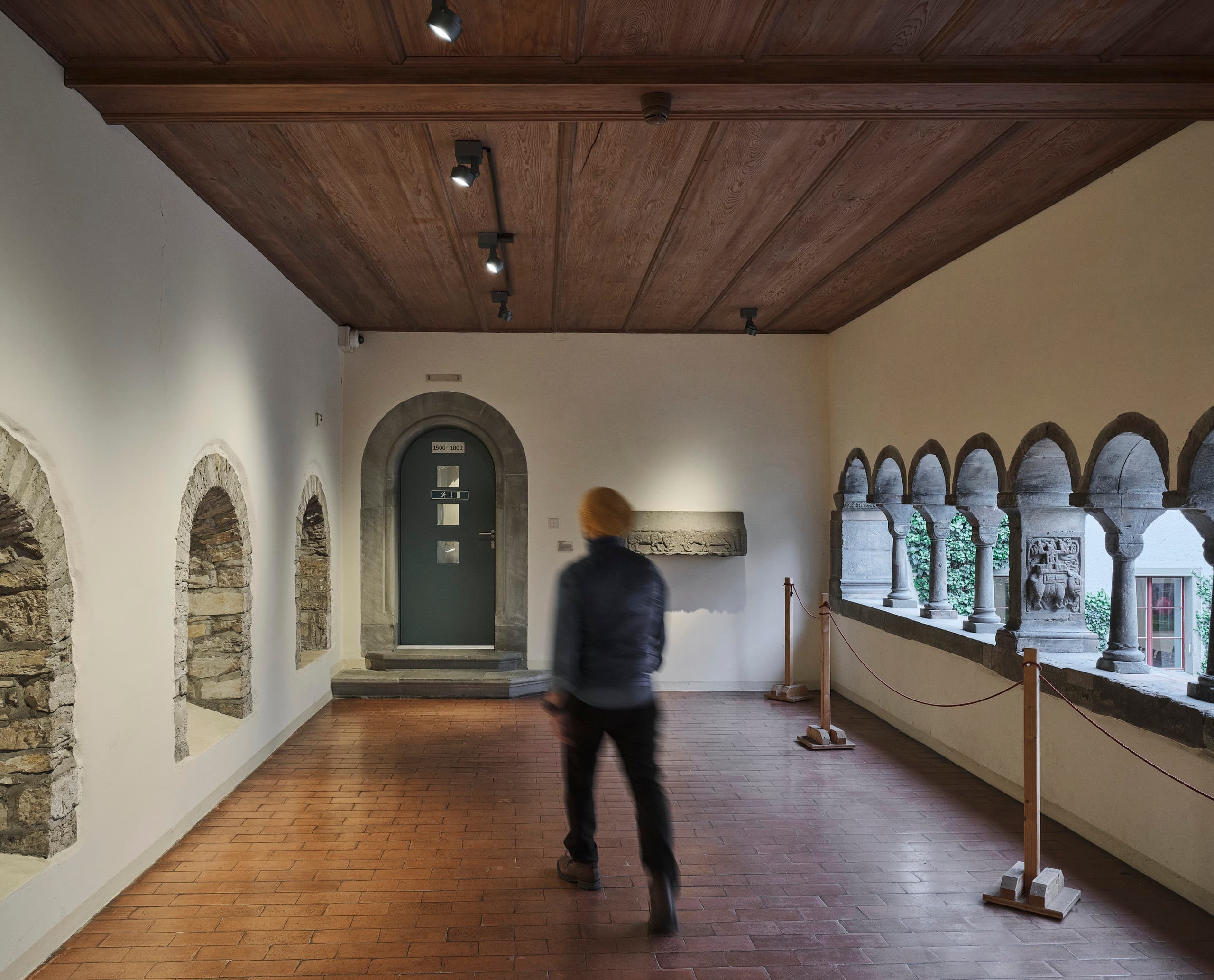 Gang in einem Museum mit runden Steinfenstern und Mann, der sich durch Raum bewegt.