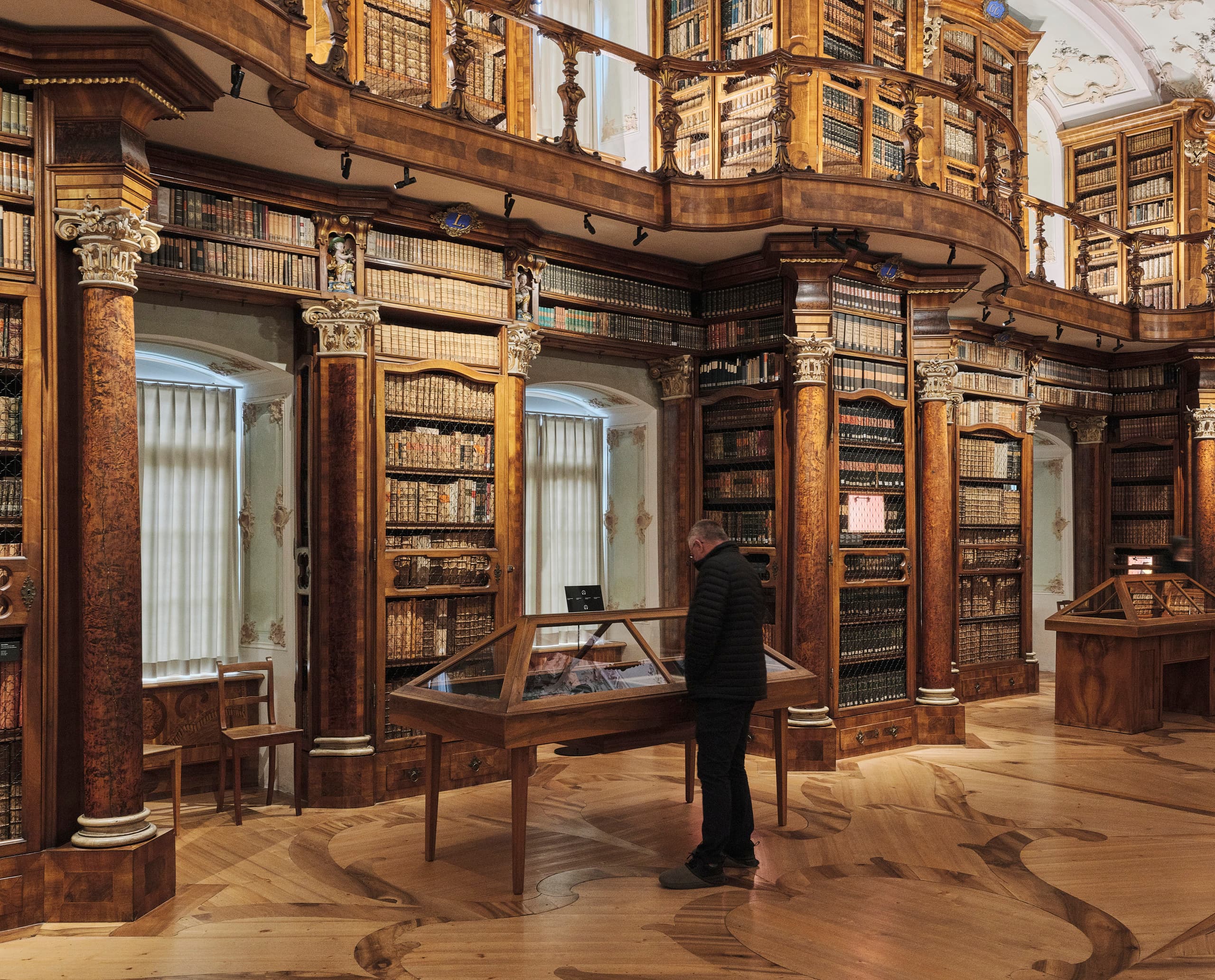 Historischer Bibliothekssaal mit hohen, reich verzierten Holzregalen, einer kunstvoll bemalten Decke und einer Person, die eine Ausstellungsvitrine betrachtet.