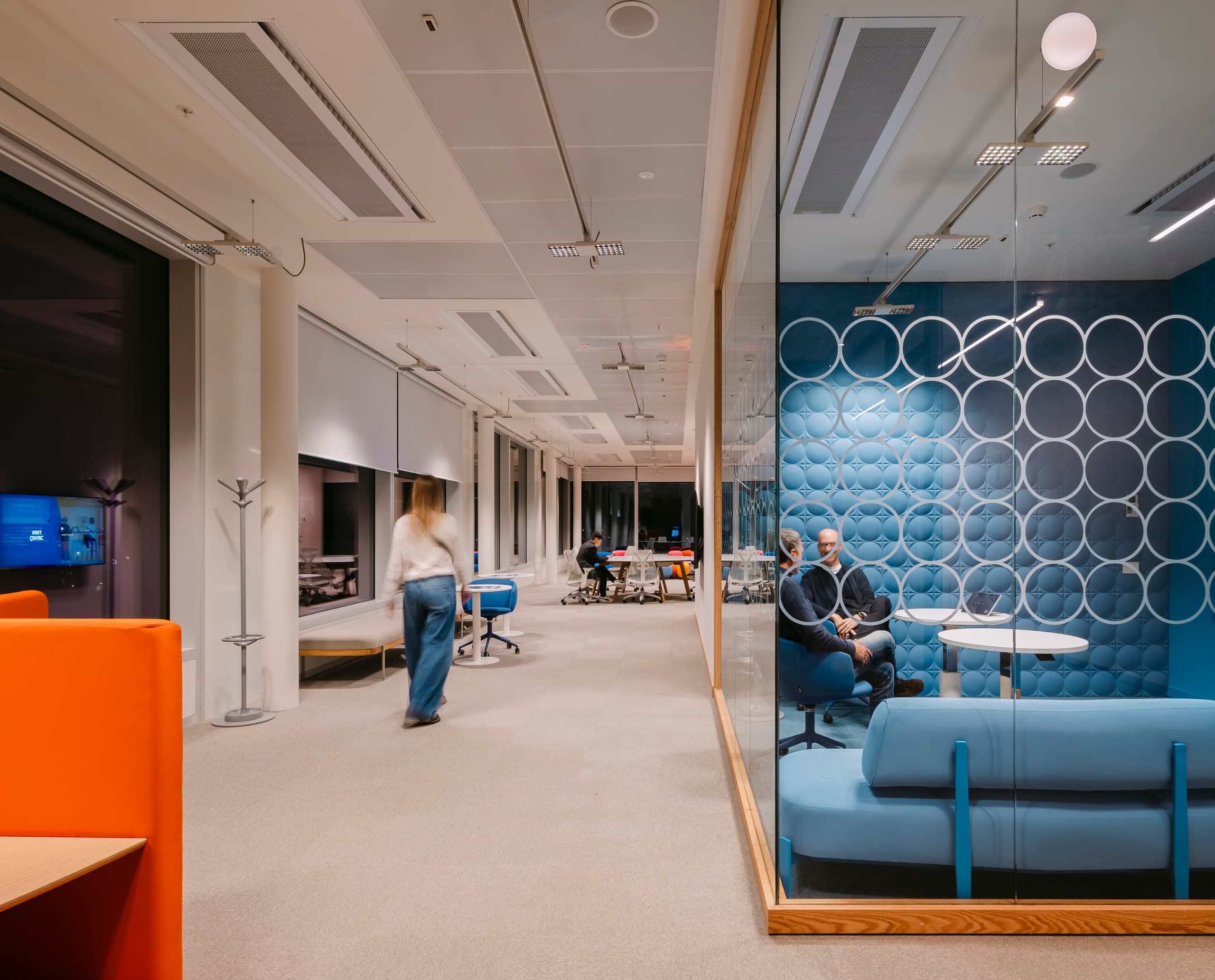 Modernes Büro mit offenem Arbeitsbereich und einem verglasten Meetingraum mit blauer Polsterwand; mehrere Personen sitzen oder bewegen sich durch den Raum, der hell beleuchtet ist.