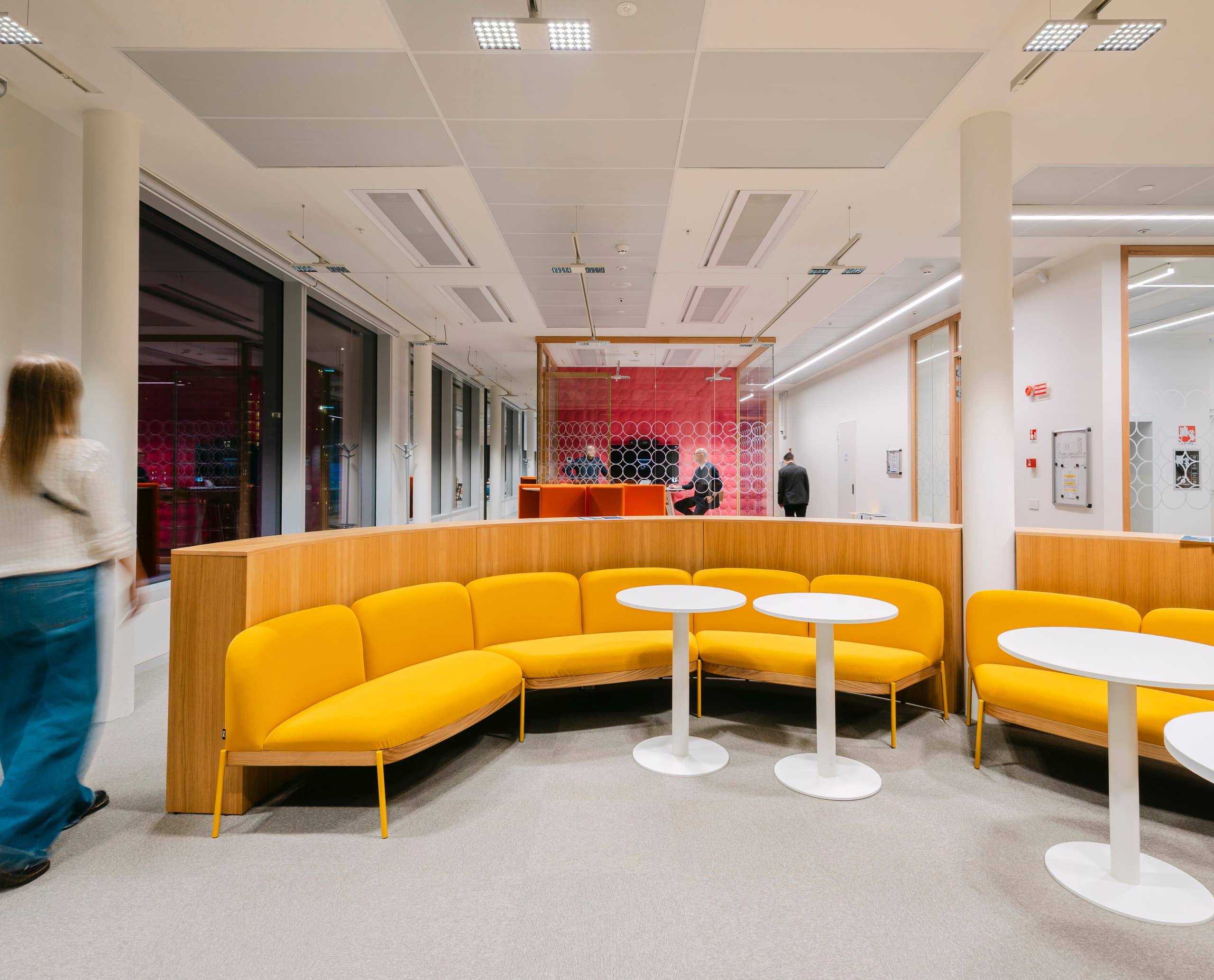 Modern eingerichteter Bürobereich mit gebogener gelber Sitzbank und weißen runden Tischen im Vordergrund; im Hintergrund verglaster Meetingraum mit roter Wandverkleidung und Personen, die sich im Raum bewegen.