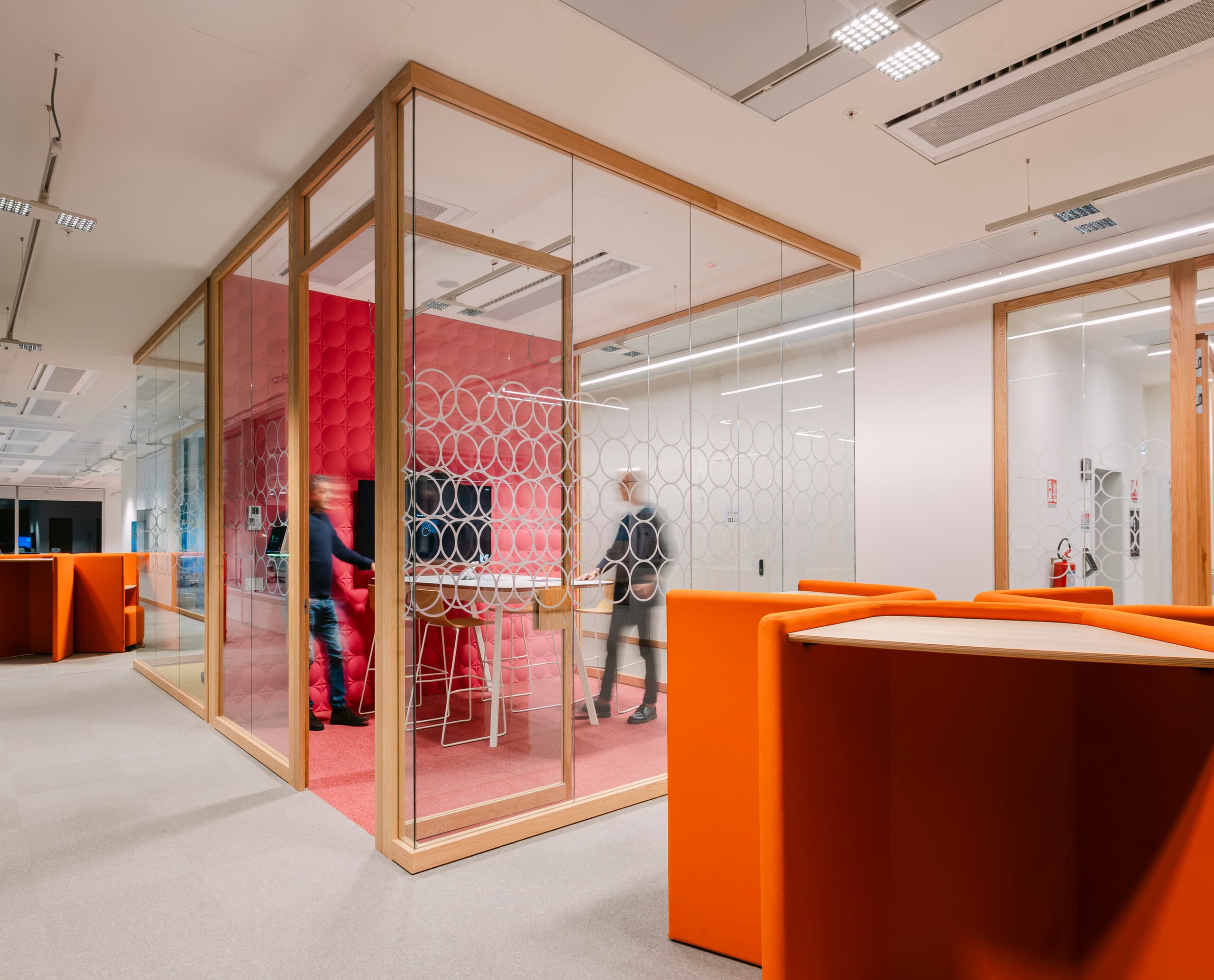 Moderner Bürobereich mit verglastem Meetingraum, dessen Wände ein kreisförmiges Muster tragen; im Inneren stehen Personen an einem hohen Tisch, während im Vordergrund orangefarbene Steh‑Arbeitsplätze zu sehen sind.