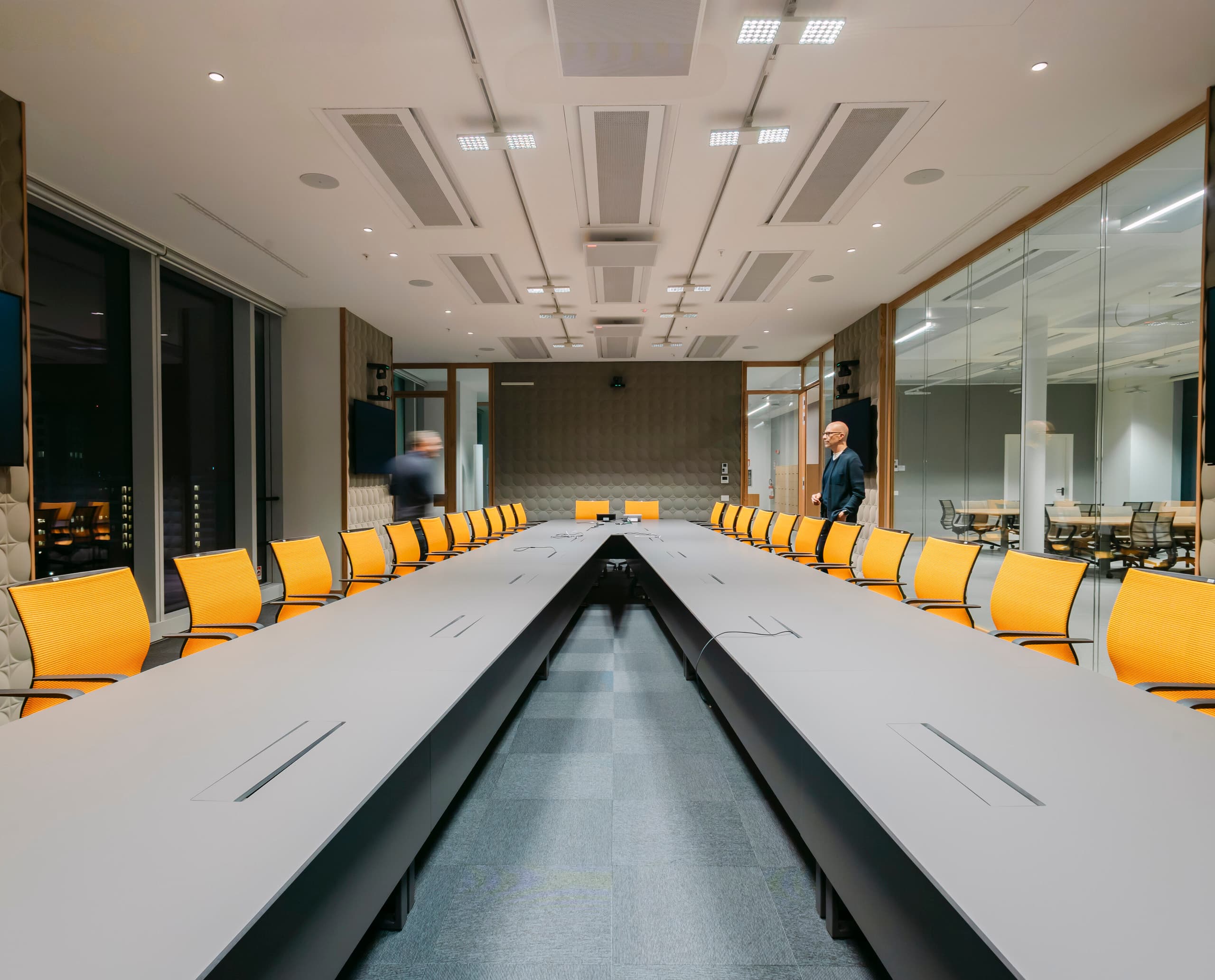 Großer, modern gestalteter Konferenzraum mit U‑förmigem Tisch und vielen gelben Drehstühlen; an den Seiten befinden sich große Bildschirme, und zwei Personen bewegen sich durch den Raum.
