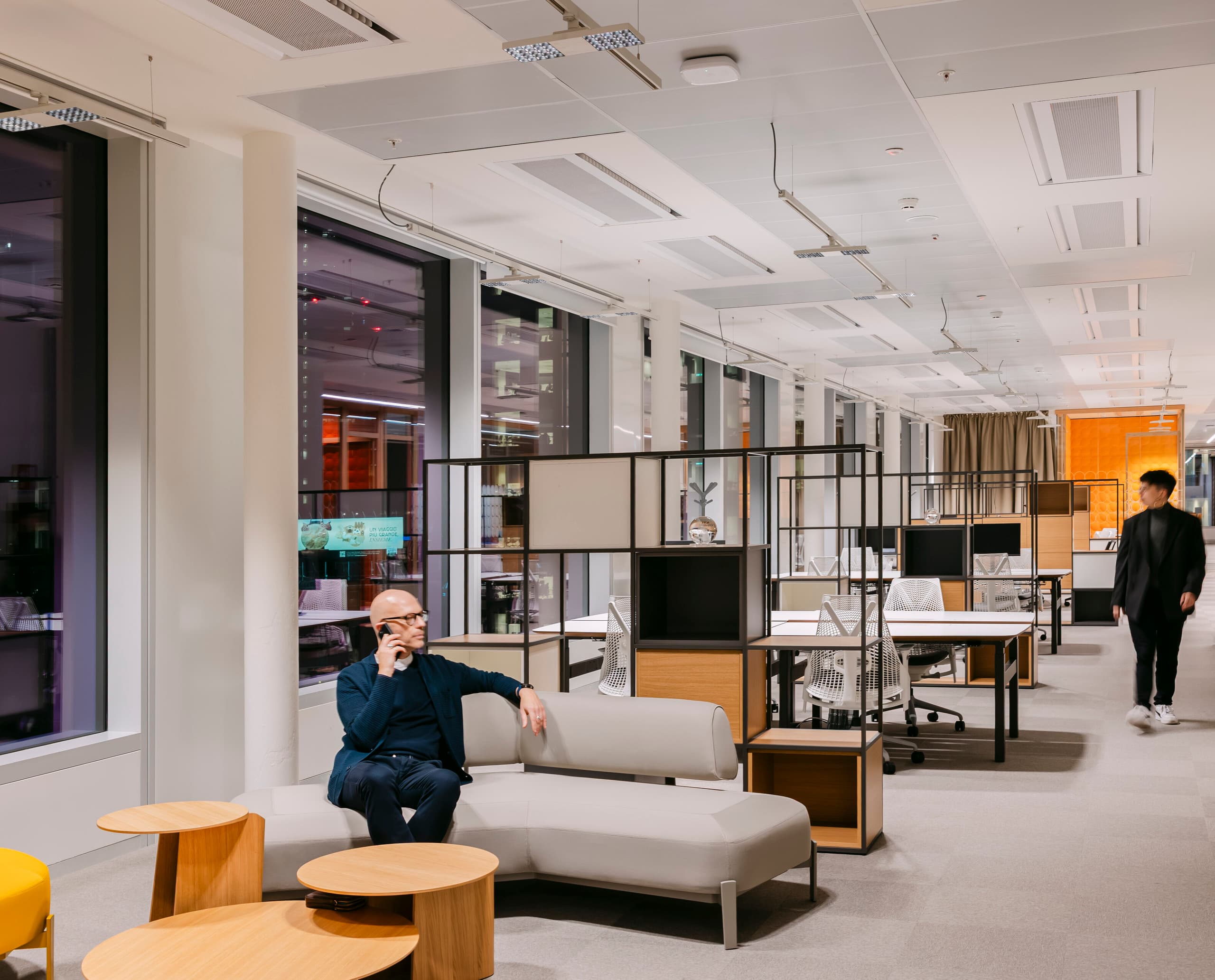 Moderner Bürobereich mit bodentiefen Fenstern, hellen Arbeitsplätzen und einem Lounge‑Sofa im Vordergrund; im Hintergrund sind mehrere Schreibtische mit Monitoren zu sehen, während eine Person durch den Raum geht.