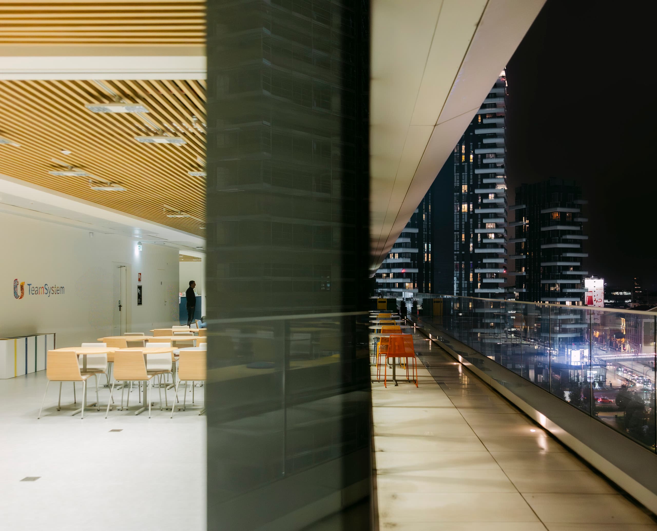 Innenbereich eines modernen Bürogebäudes mit Holzlamellendecke und hellen Sitzbereichen auf der linken Seite; rechts eine breite, überdachte Terrasse mit Glasbalustrade und Blick auf beleuchtete Hochhäuser bei Nacht.