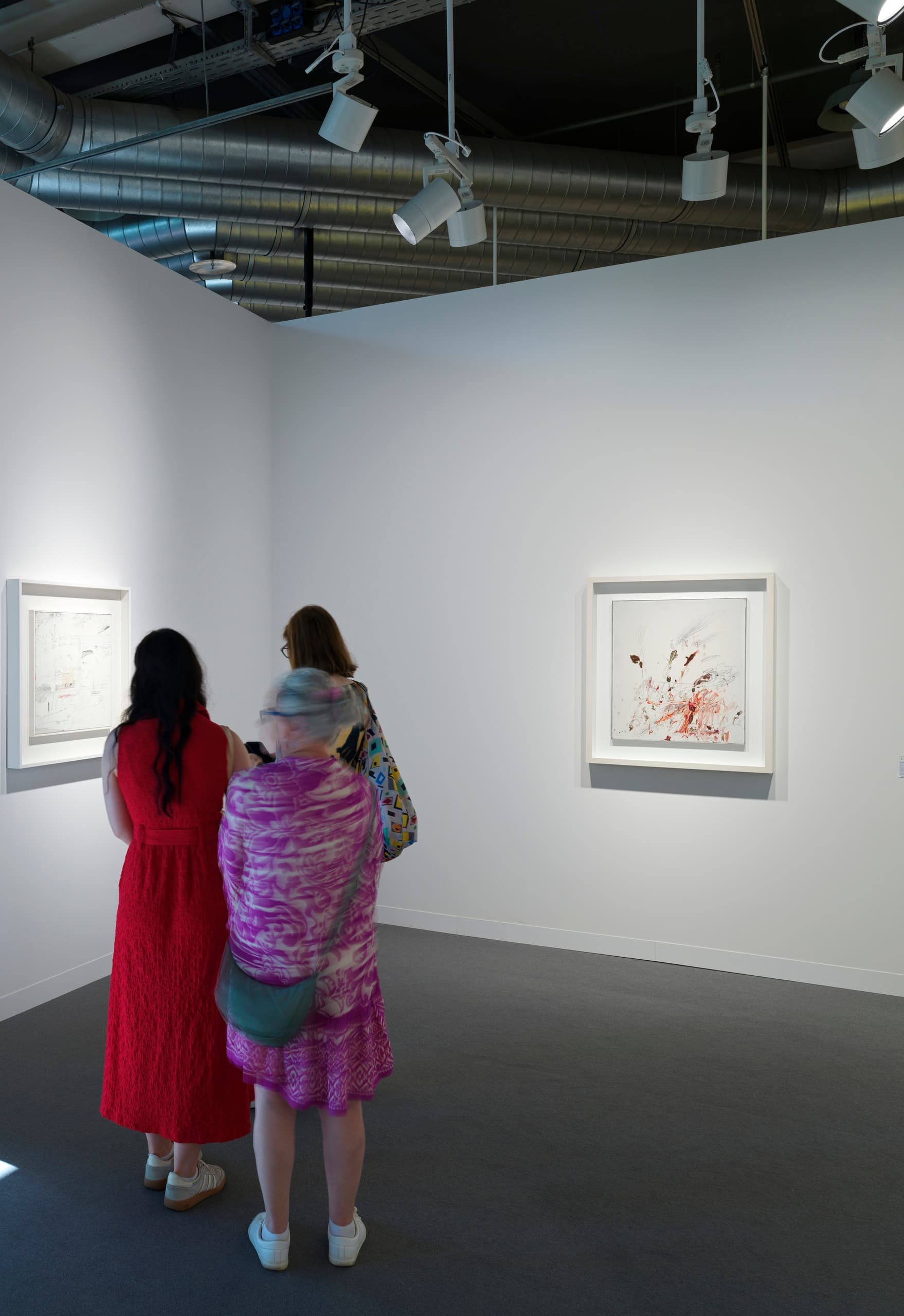 Drei Personen schauen sich Kunst an einer weißen Wand auf einem Messestand der Art Basel an.