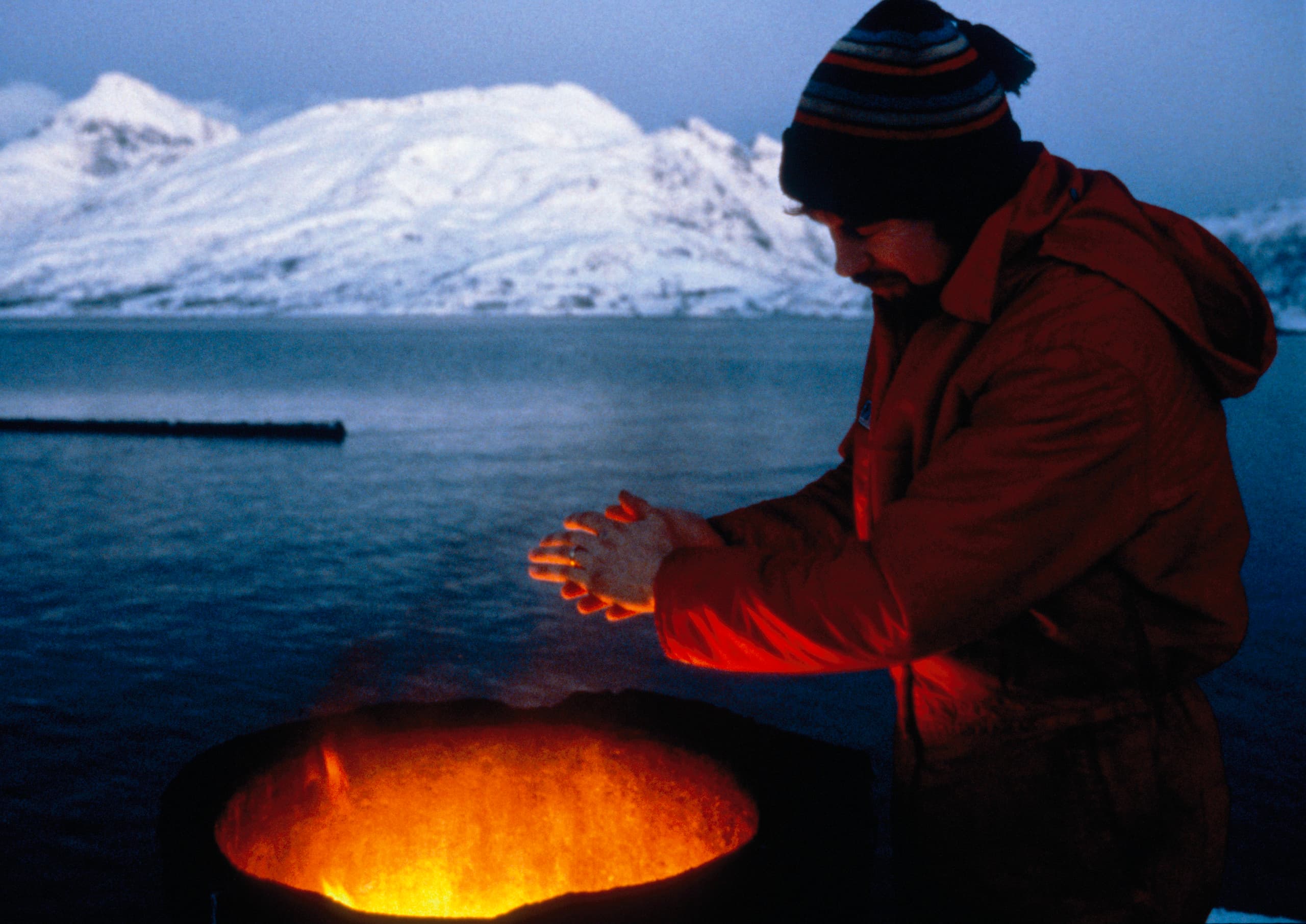 Mann wärmt sich am Feuer vor verschneiter Bergkulisse, Lichtdesign-Konzept.