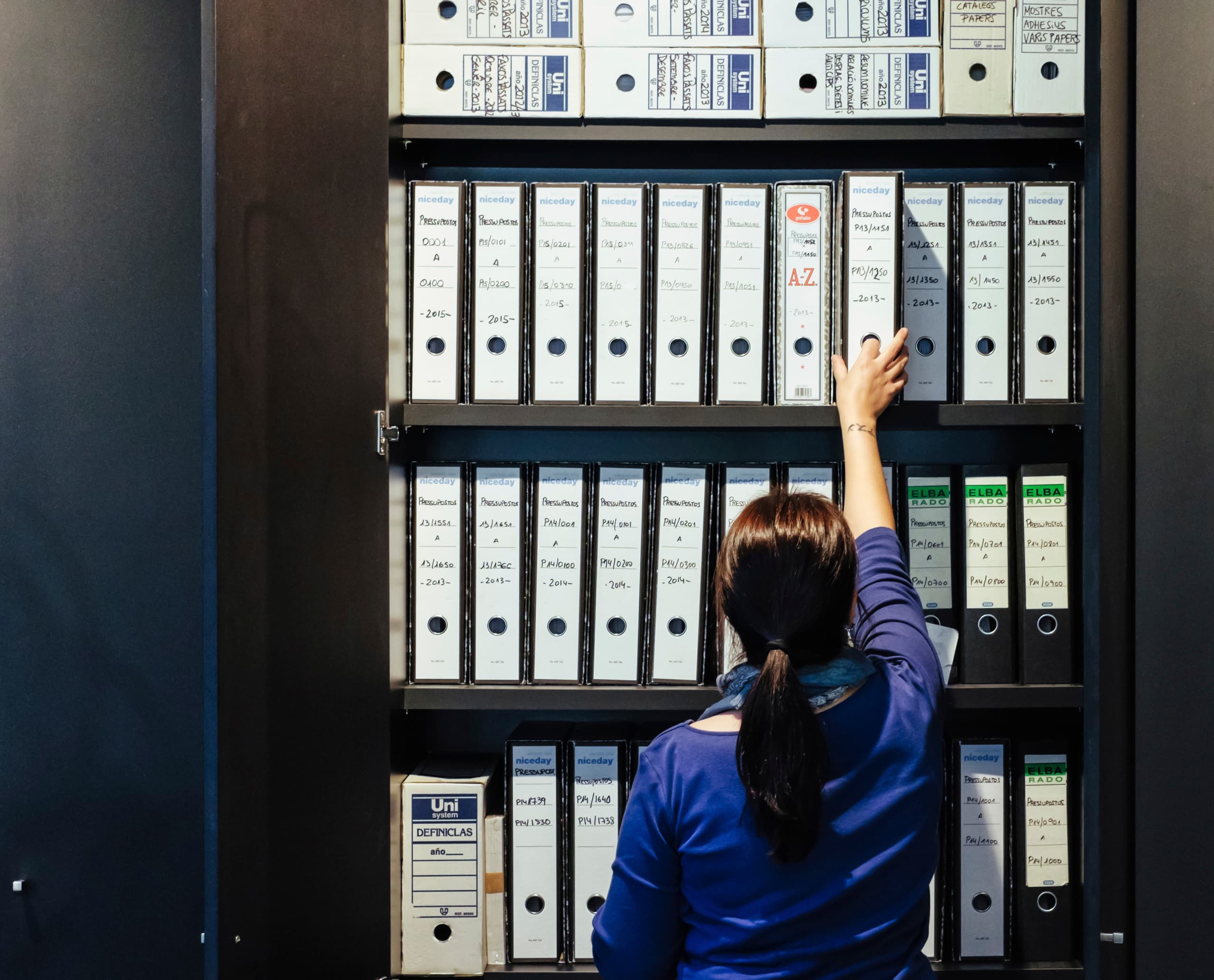 Frau greift nach Ordnern im Büro von Graficas Varias, beleuchtet mit ERCO LED für ergonomische Arbeitsplatzbeleuchtung.