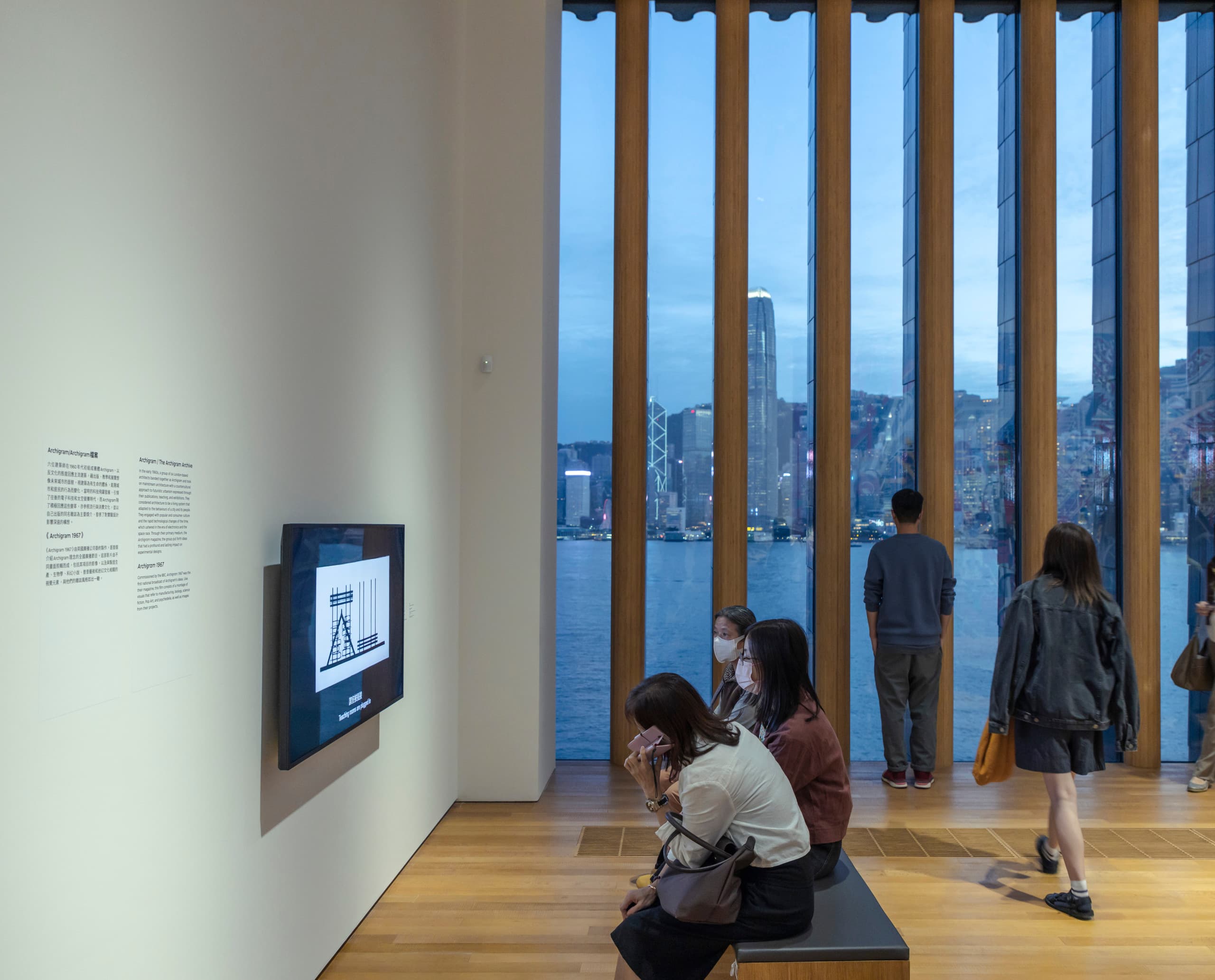 Besucher im M+ Museum betrachten Kunstwerke, beleuchtet mit ERCO Strahlern, mit Blick auf Hongkongs Skyline.