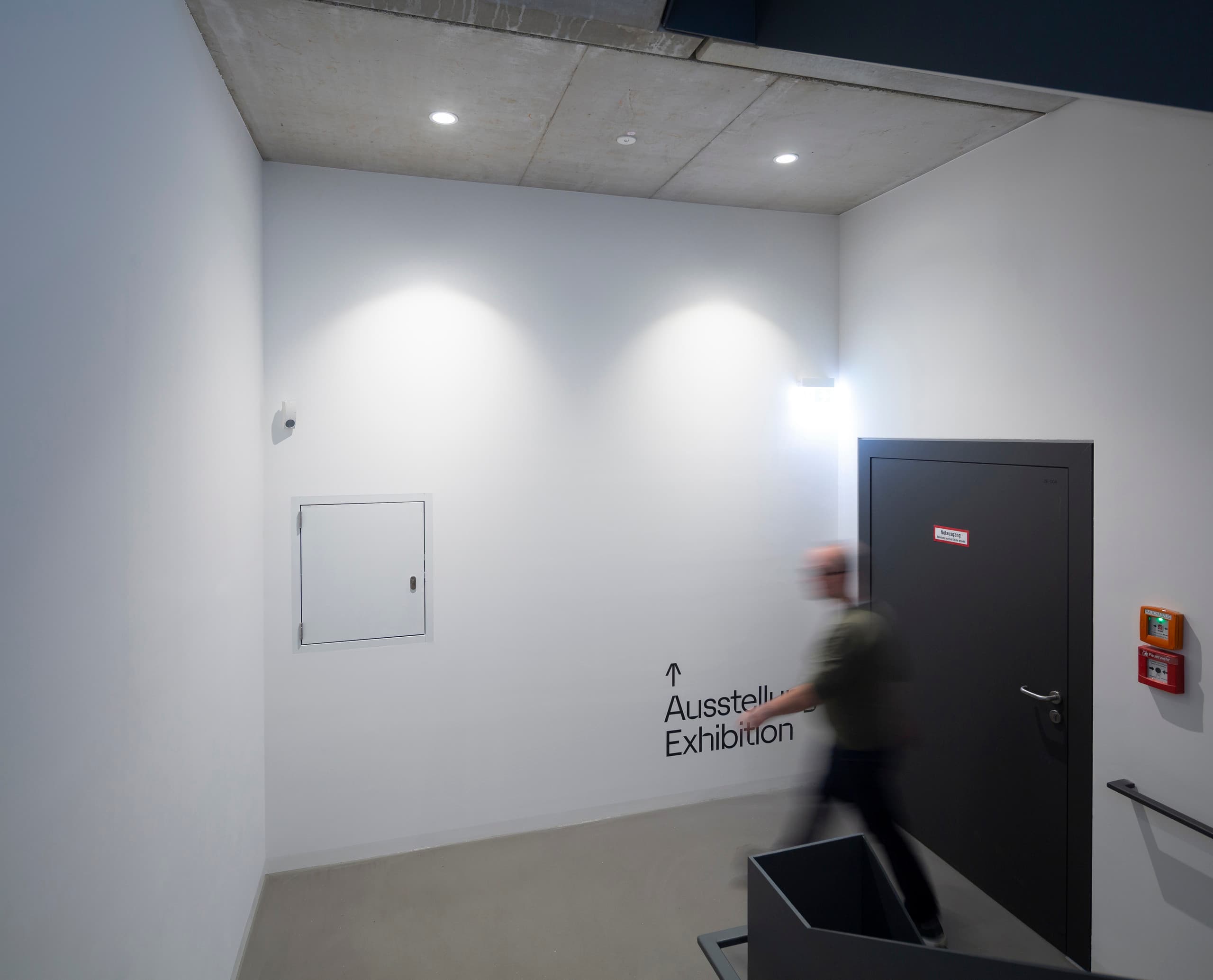 ERCO LED-Beleuchtung im Blockhaus Dresden: Flur mit Hinweisschild zur Ausstellung und architektonischer Beleuchtung.
