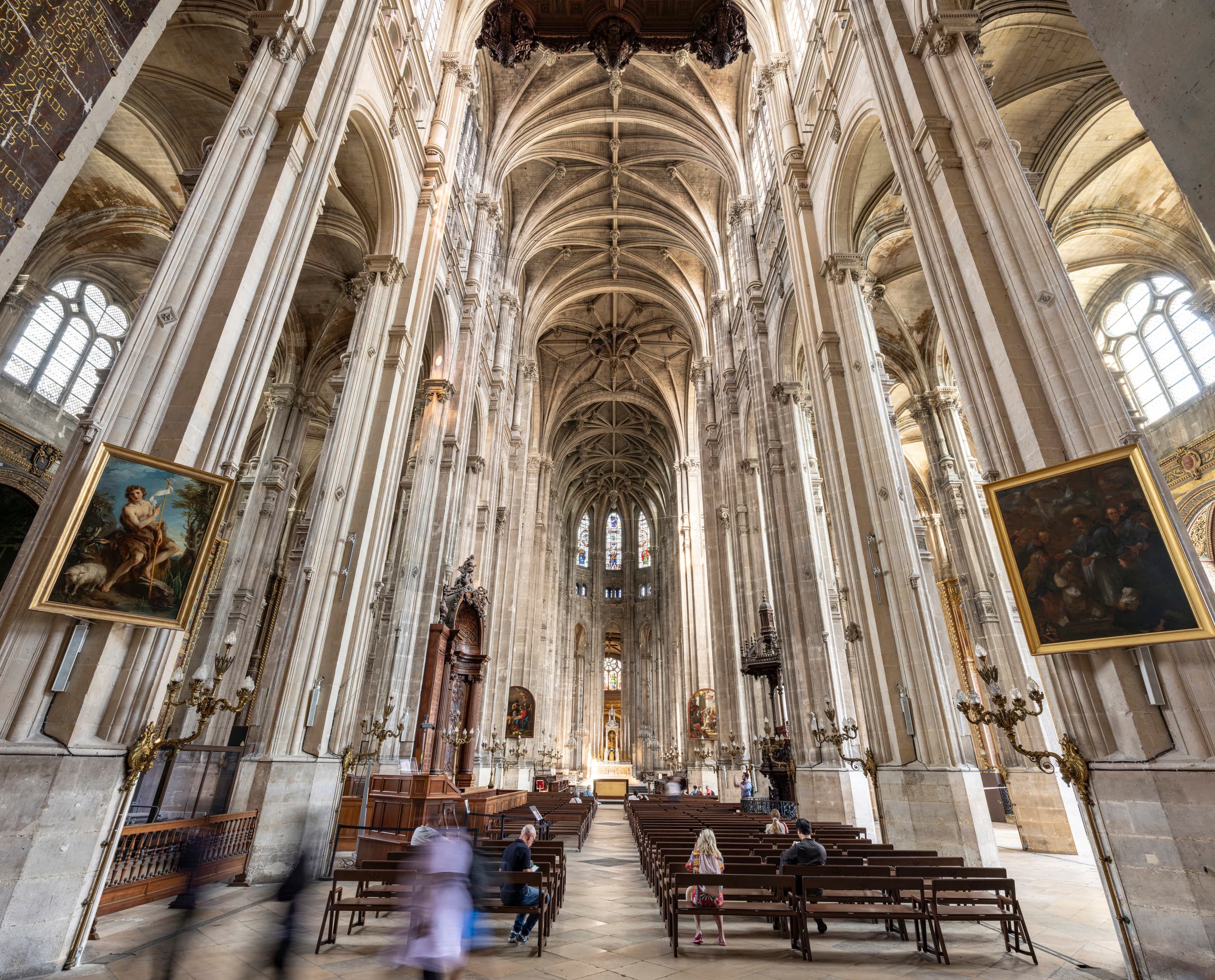 Die Weitwinkelaufnahme zeigt den Innenraum einer großen gotischen Kirche mit hohen Säulen aus hellem Stein, Gewölbedecken sowie zwei gerahmten Gemälden, die seitlich hängen. Im Vordergrund stehen Reihen von Holzbänken. Mehrere Besucher sitzen auf den Bänken oder gehen umher. Der Mittelgang führt zu dem etwas entfernten, beleuchteten Altar, der von kunstvollen architektonischen Details und Buntglasfenstern eingerahmt wird.