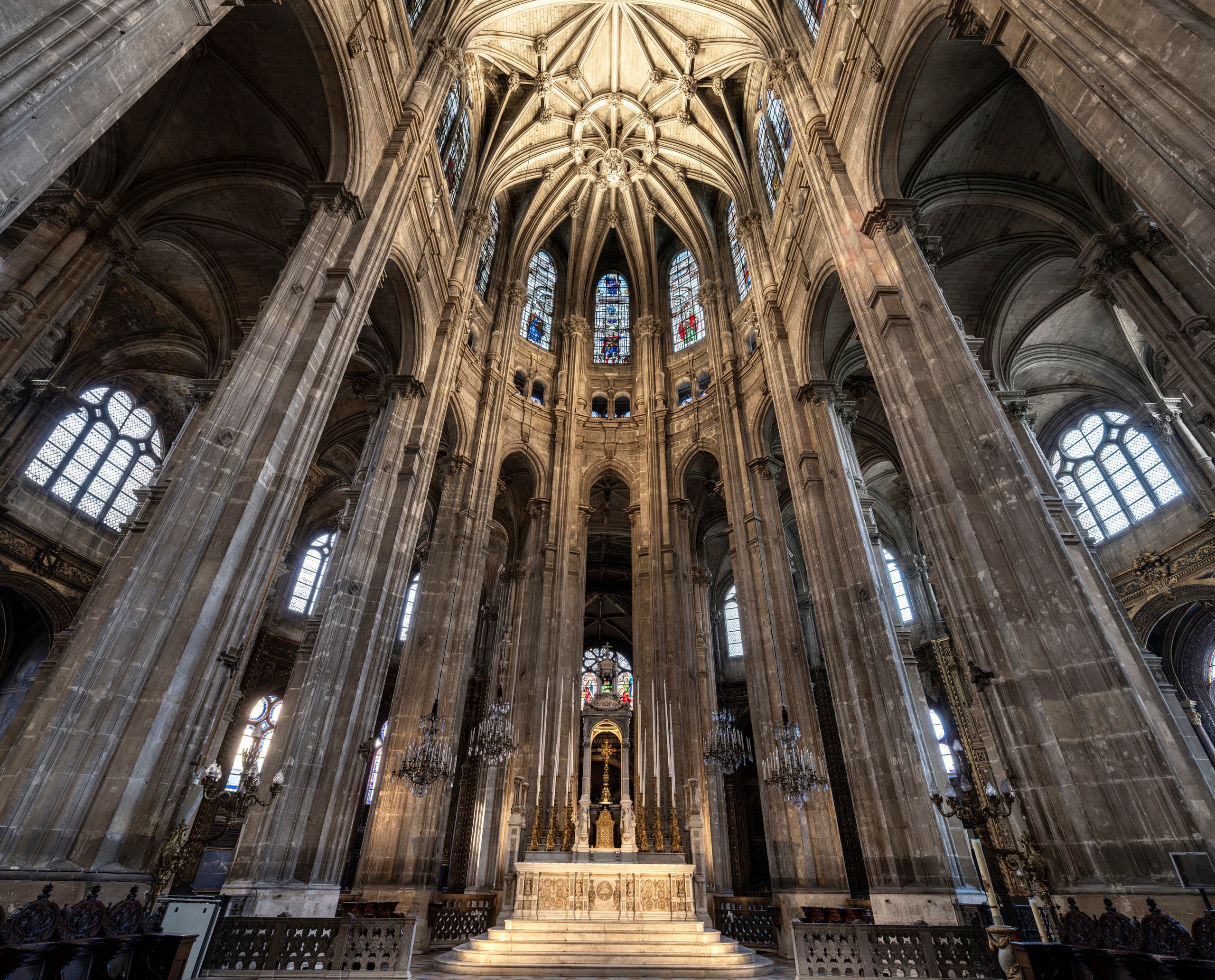 Der prächtige Chorraum einer großen, historischen Kirche ist zu sehen: hohe, schlanke Steinsäulen, reich verzierte Gewölbe und Buntglasfenster. Im Mittelpunkt des Bildes steht der erhöhte Altar aus hellem Stein, umgeben von kunstvollen Skulpturen und Kerzenleuchtern. Durch die hohen Fenster strömt Licht herein und setzt die monumentale Architektur effektvoll in Szene.