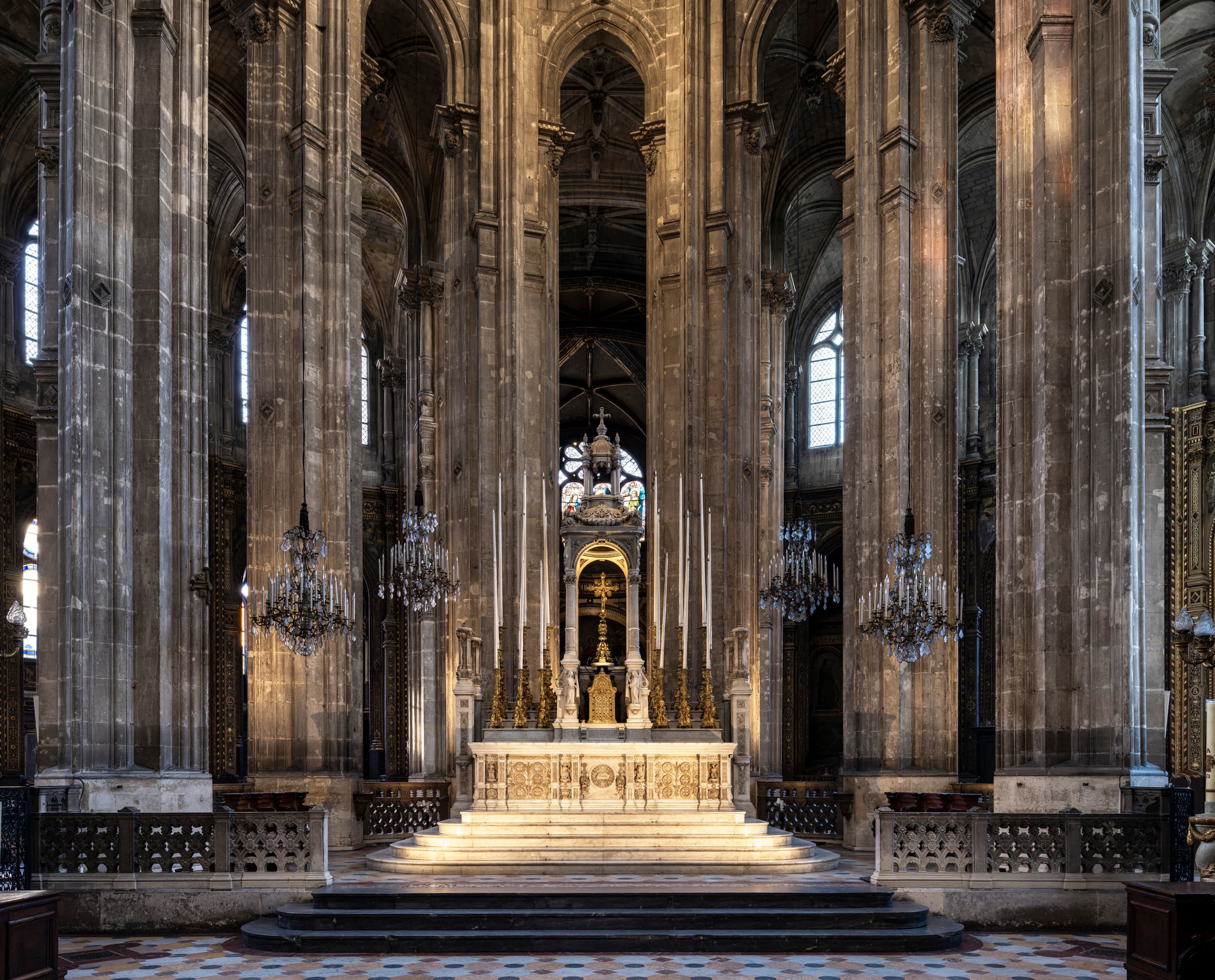 Vorderansicht eines prächtigen Kirchenaltars, der von hohen Steinsäulen, kunstvollen Kronleuchtern und aufwendigen gotischen architektonischen Details eingerahmt ist. In der Mitte, unter hohen Gewölbebögen und großzügigen Bogenfenstern, steht der beleuchtete Altar mit seinen geschnitzten Verzierungen und großen Kerzenleuchtern.