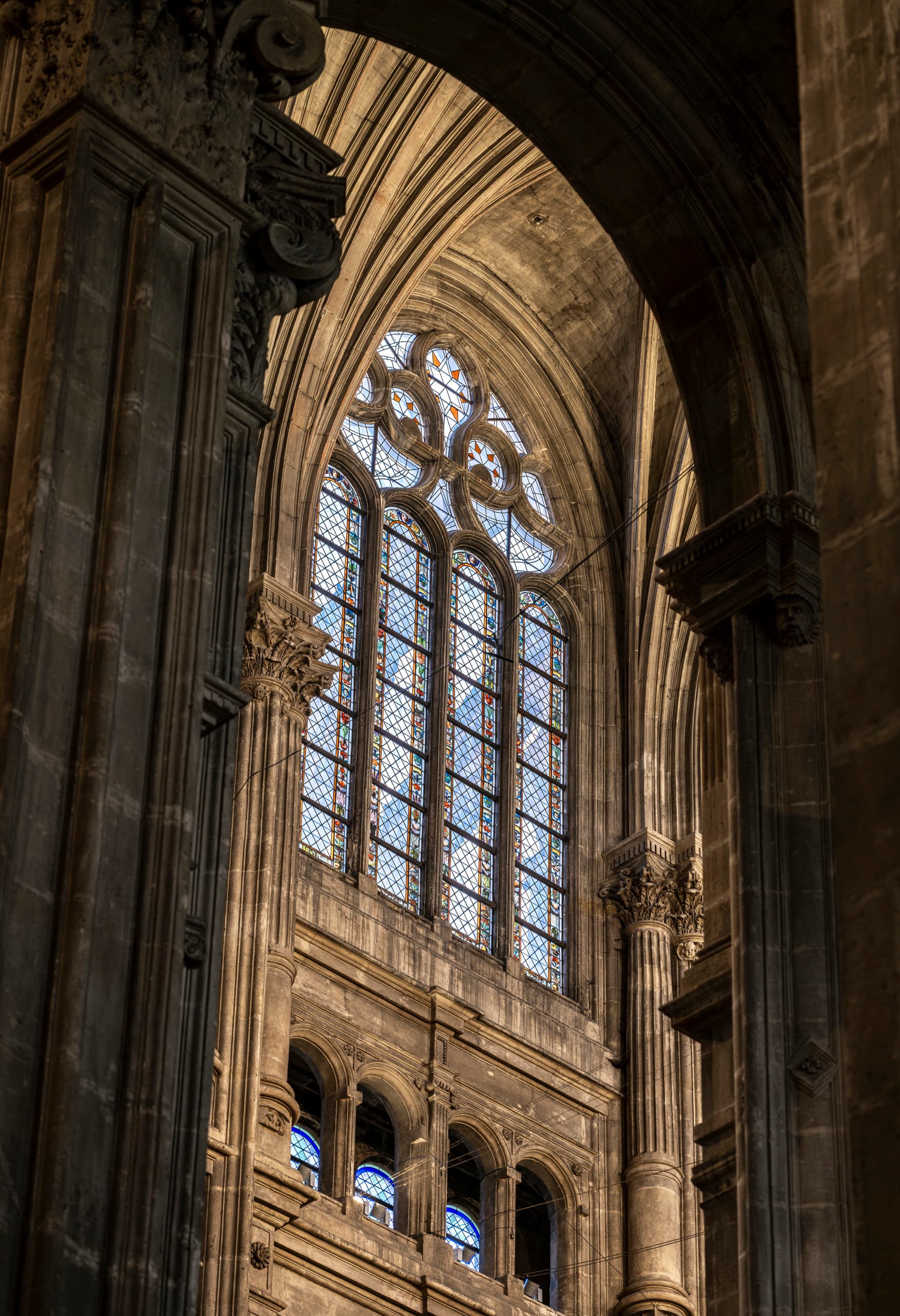 Nahaufnahme eines hohen gotischen Kirchenfensters mit aufwendigem Steinmaßwerk und mehreren Buntglasfenstern. Das Fenster wird von großen Steinsäulen und kunstvollen architektonischen Schnitzereien eingerahmt und sanft von natürlichem Licht erhellt.