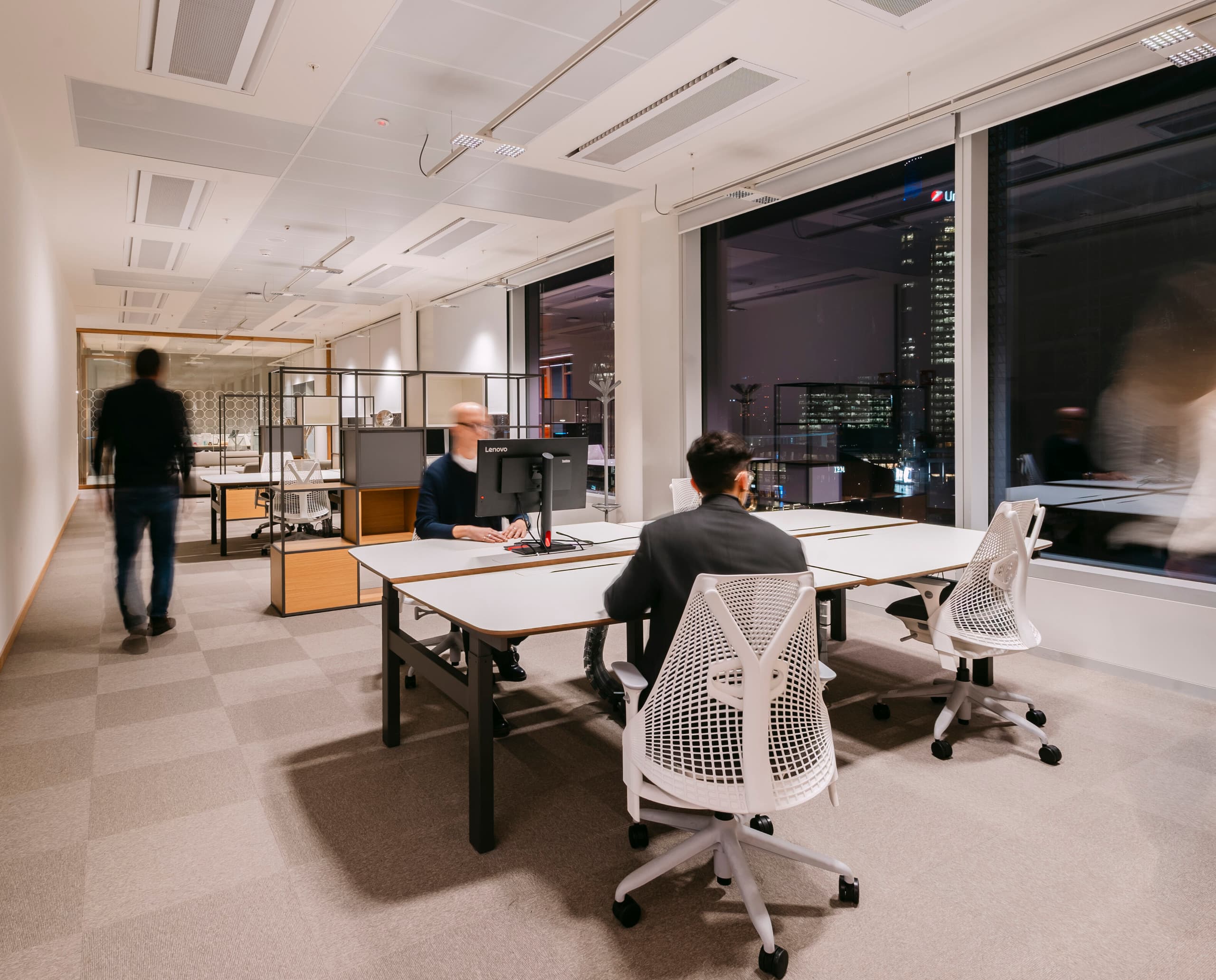 Heller, modern eingerichteter Bürobereich mit mehreren Schreibtischen und ergonomischen Bürostühlen; eine Person sitzt an einem Arbeitsplatz vor einem Bildschirm, während zwei weitere Personen durch den Raum gehen, und große Fenster geben den Blick auf eine nächtliche Stadt frei.