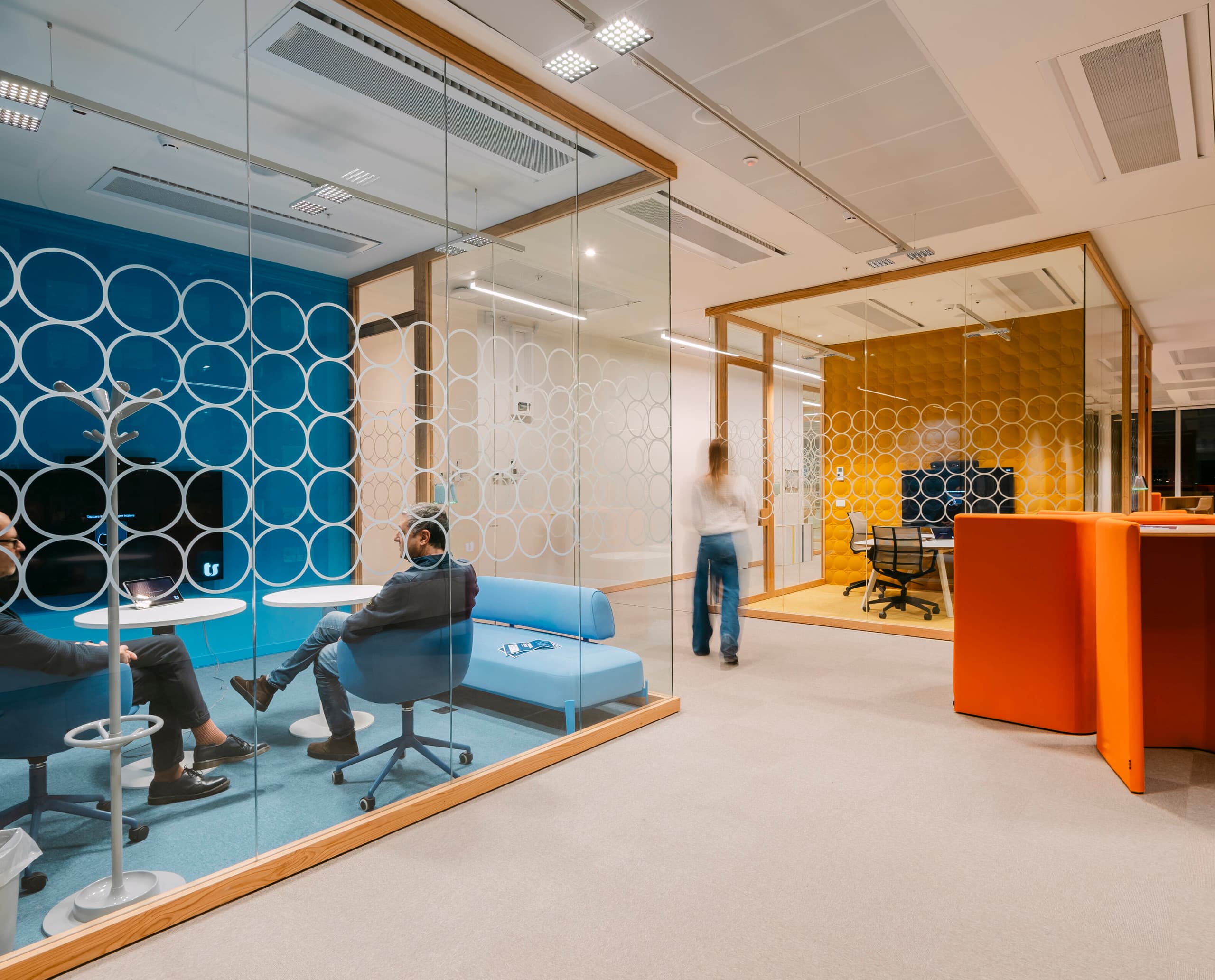 Moderner Bürobereich mit zwei verglasten Meetingräumen, einem blauen und einem gelben Raum mit kreisförmigem Muster auf den Glaswänden; im vorderen Meetingraum sitzen zwei Personen auf blauen Sesseln, im hinteren Raum steht ein großer Tisch, und eine weitere Person geht den Flur entlang.