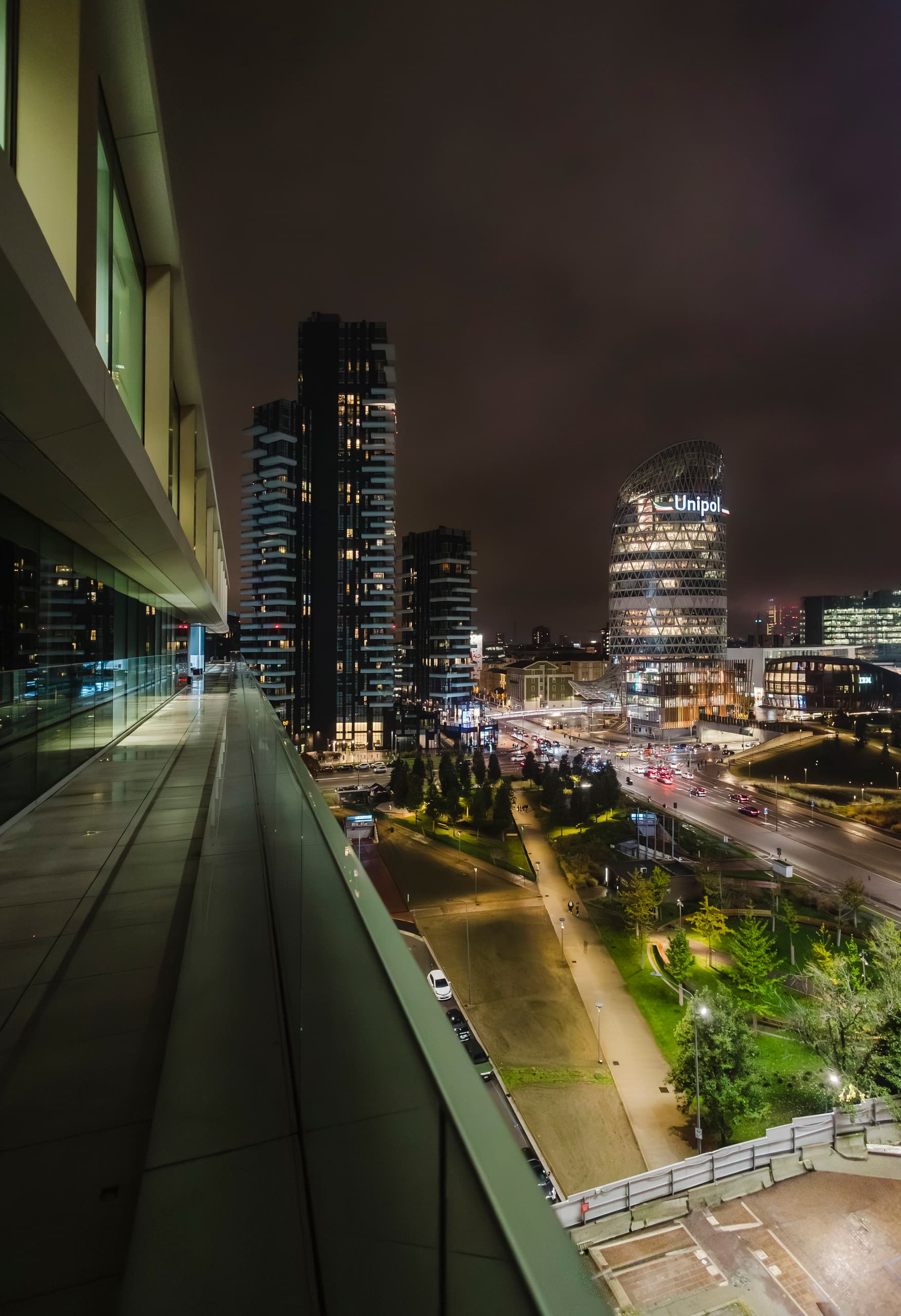 Nachtaufnahme einer erhöhten Terrasse mit Glasgeländer, die den Blick auf moderne, beleuchtete Hochhäuser und eine belebte Stadtlandschaft mit Straßen und Grünflächen freigibt.