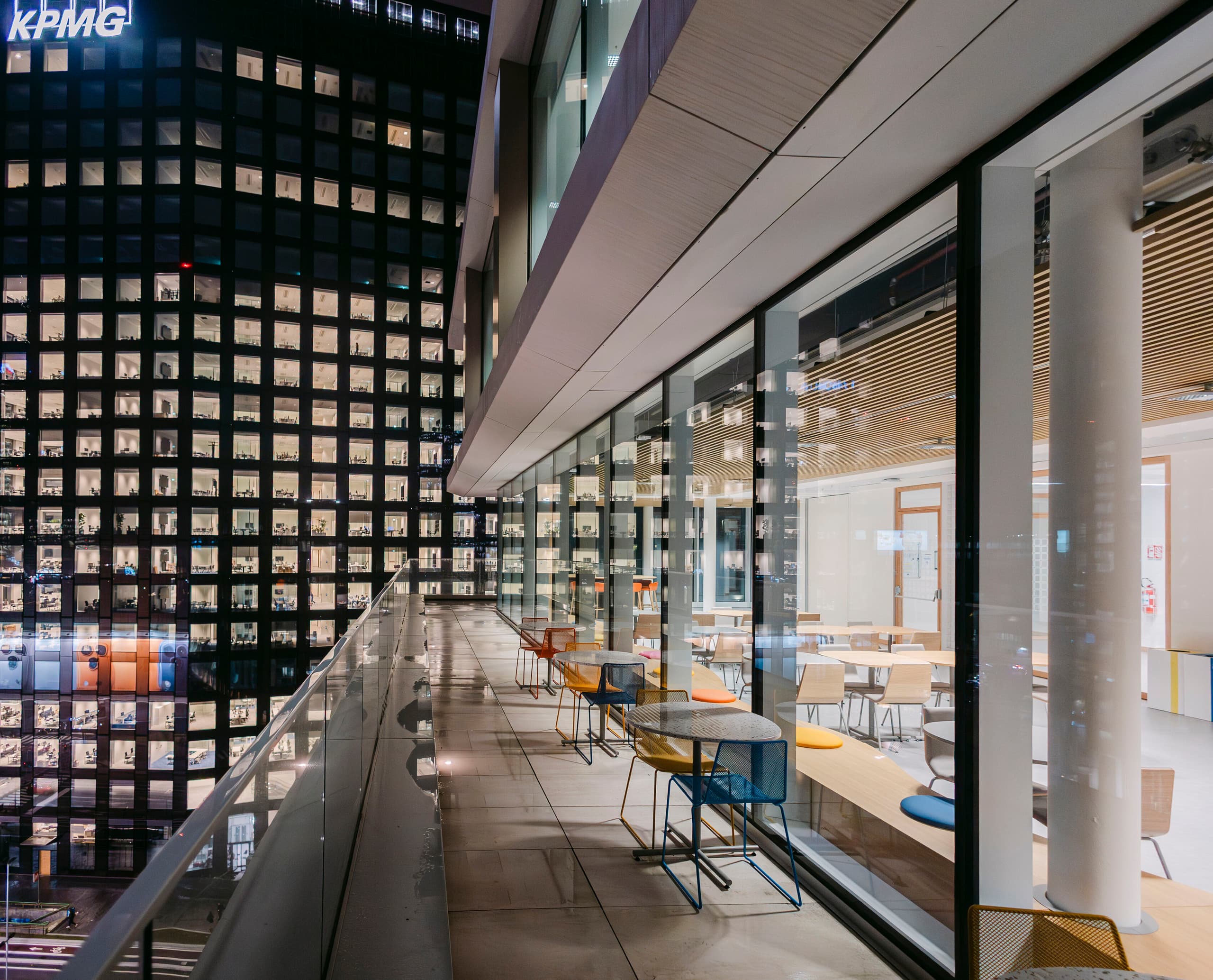 Nachtaufnahme einer modernen Büroterrasse mit Glasgeländer und farbigen Stühlen entlang der Fensterfront; rechts ist ein heller Innenbereich mit Sitzgruppen sichtbar, links steht ein hoch beleuchtetes Bürogebäude mit zahlreichen Fenstern
