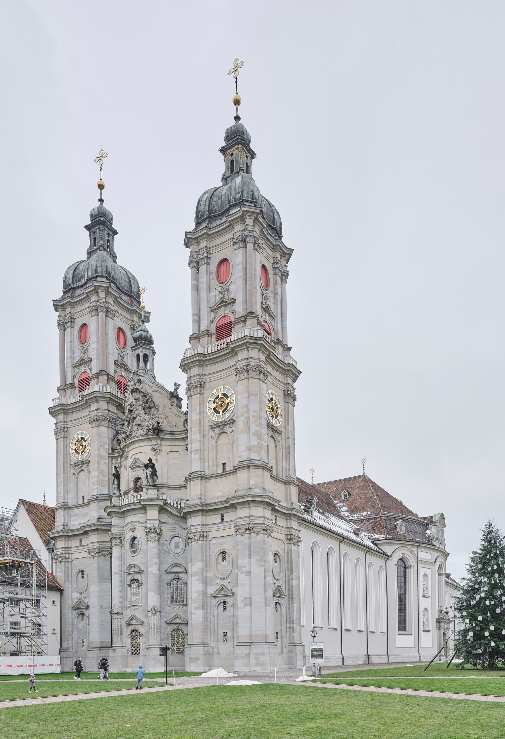 Frontansicht einer großen barocken Kirche mit zwei hohen Türmen, hellen Steinfassaden und grünem Vorplatz unter bewölktem Himmel.