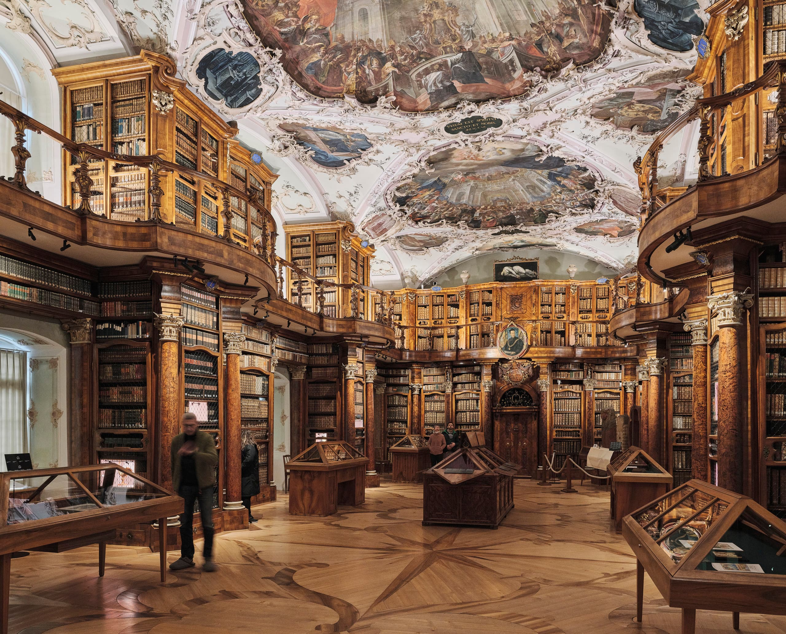 Prächtig ausgestatteter historischer Bibliothekssaal mit kunstvoll bemalter Decke, hohen Holzregalen voller Bücher und mehreren Ausstellungsvitrinen.
