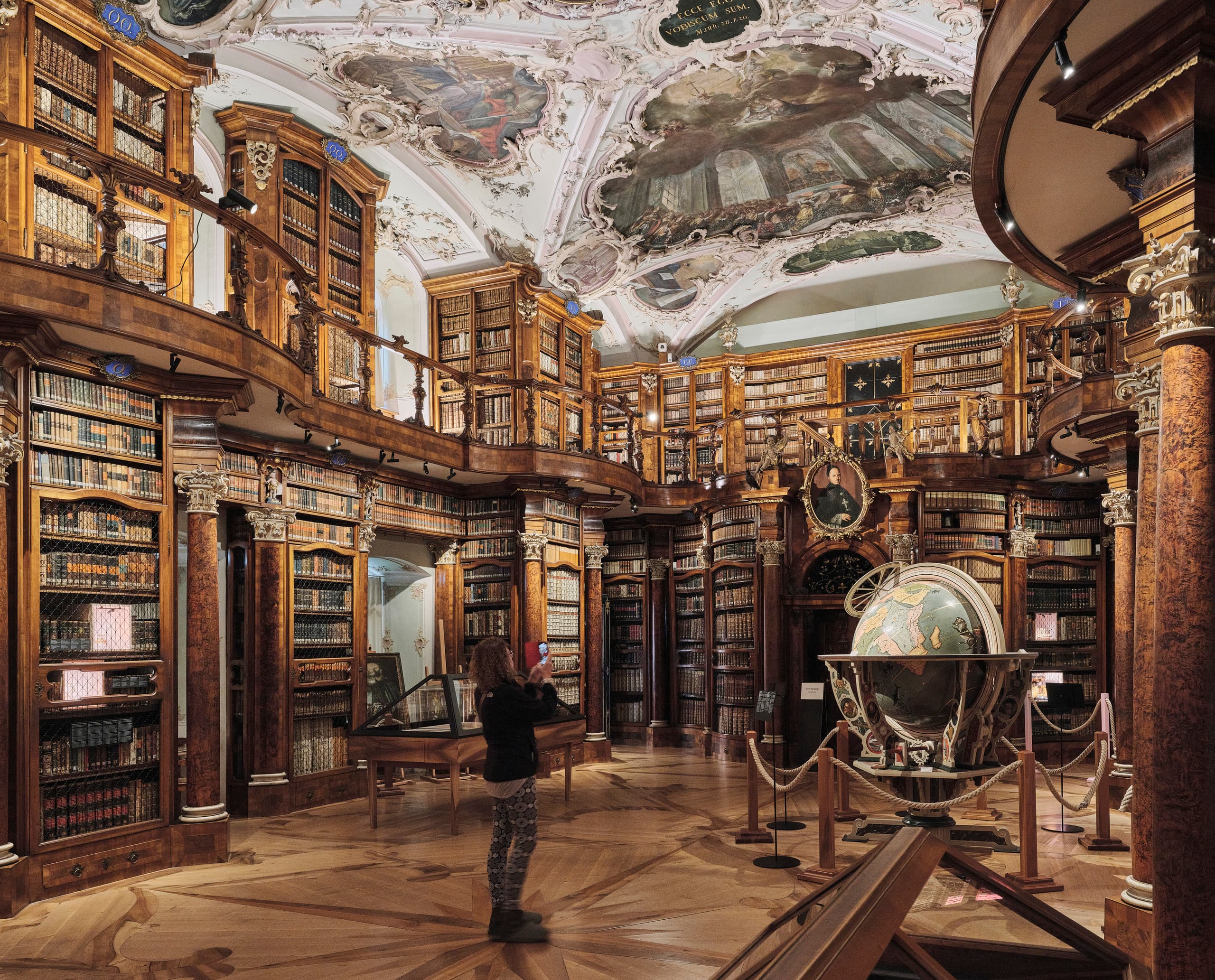 Historischer Bibliothekssaal mit kunstvoll bemalter Decke, hohen Holzregalen voller Bücher, einem großen Globus und einer Person, die ein Foto macht.