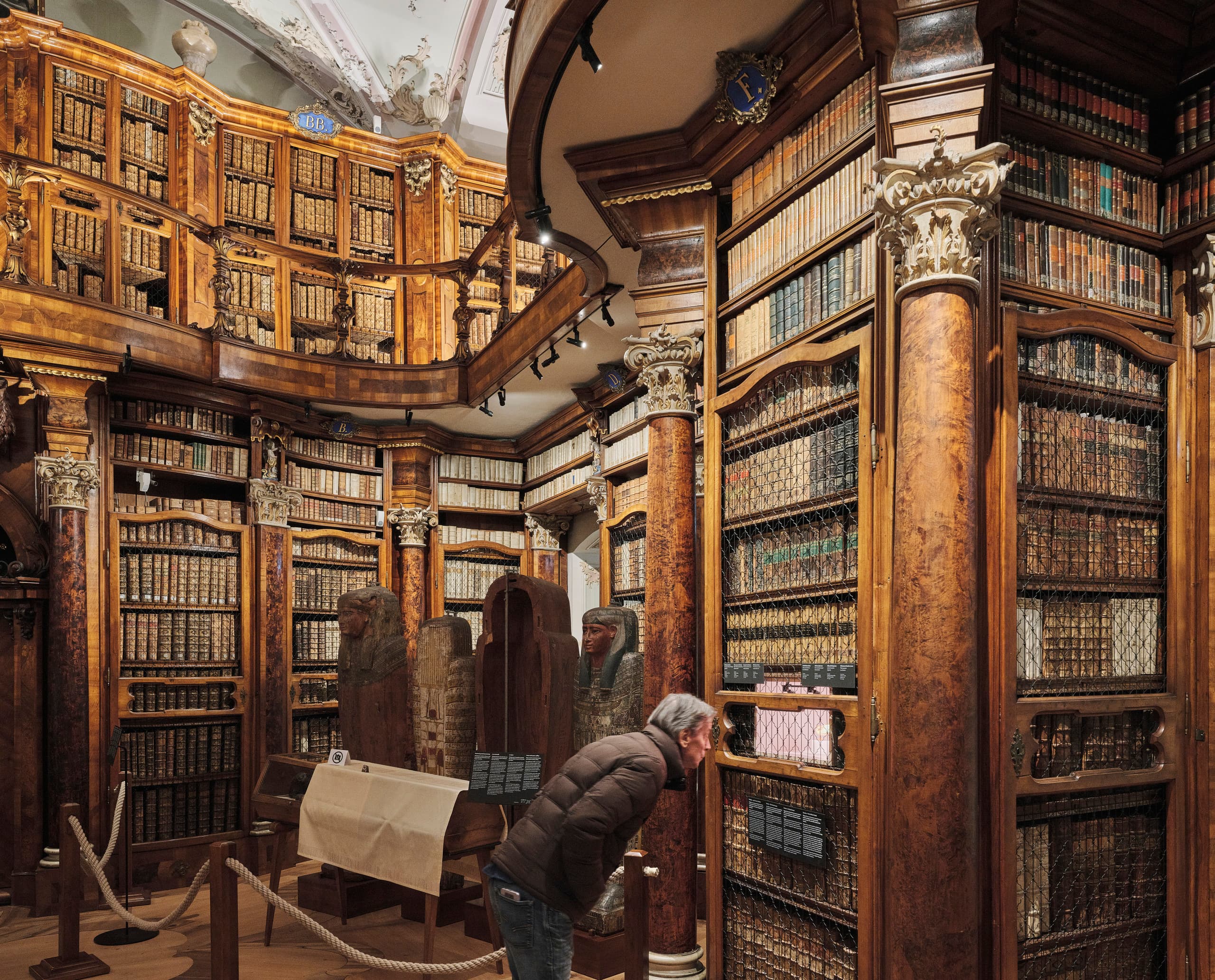 Detailansicht eines historischen Bibliothekssaals mit hohen verzierten Holzregalen und einer Person, die eine Ausstellungsvitrine betrachtet.