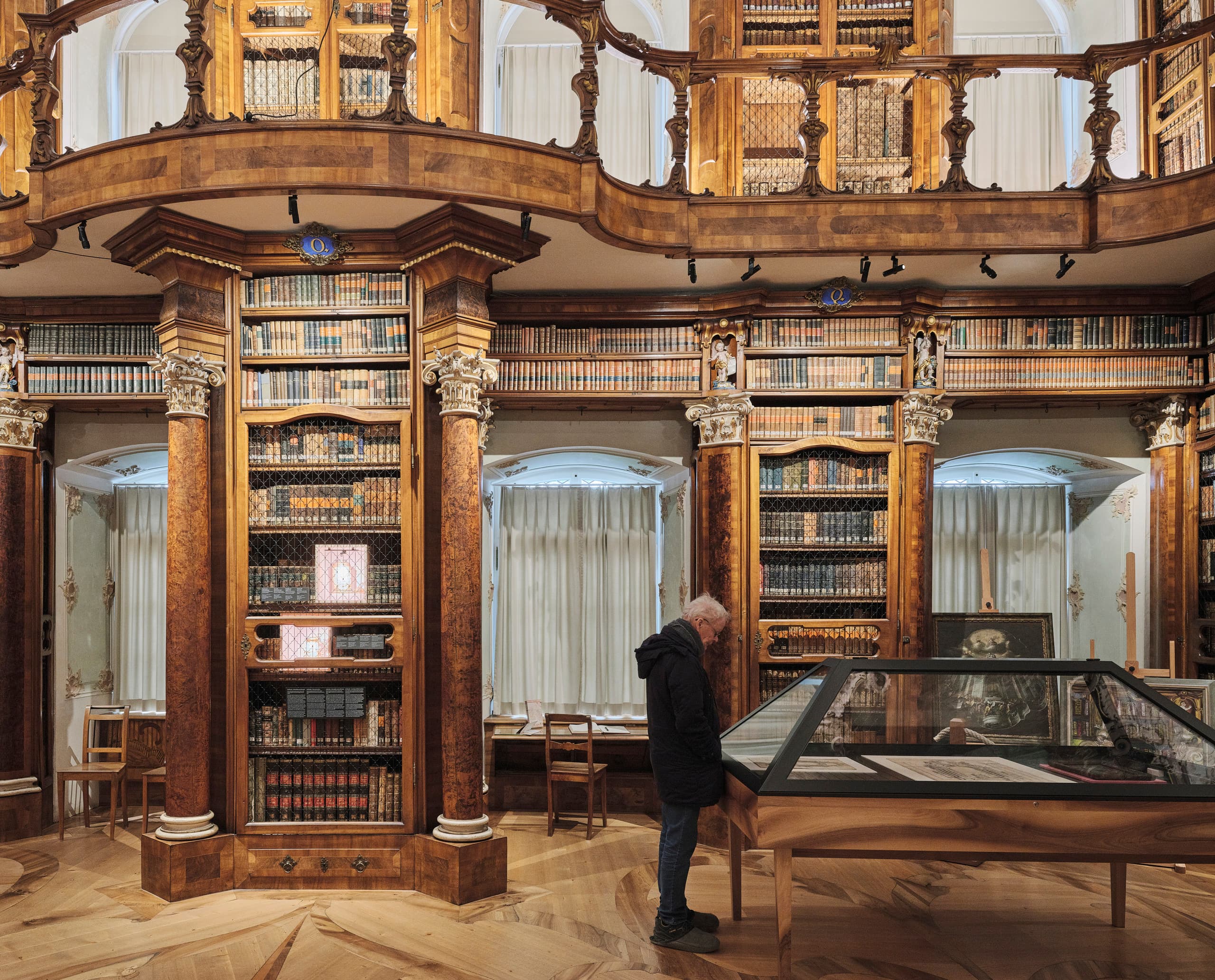 Detailansicht eines historischen Bibliothekssaals mit reich verzierten Holzregalen und einer Person, die eine Ausstellungsvitrine betrachtet.
