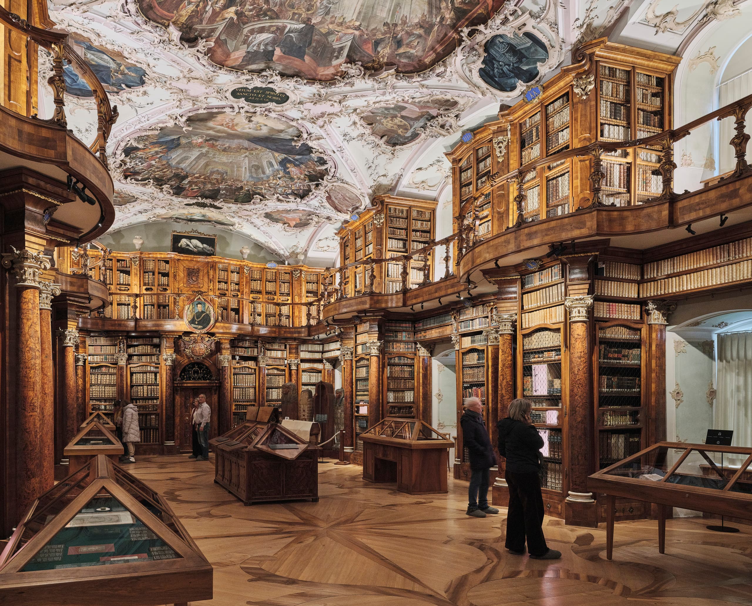 Historischer Bibliothekssaal mit reich verzierten Holzregalen, kunstvoll bemalter Decke und mehreren Personen, die Ausstellungsvitrinen betrachten.
