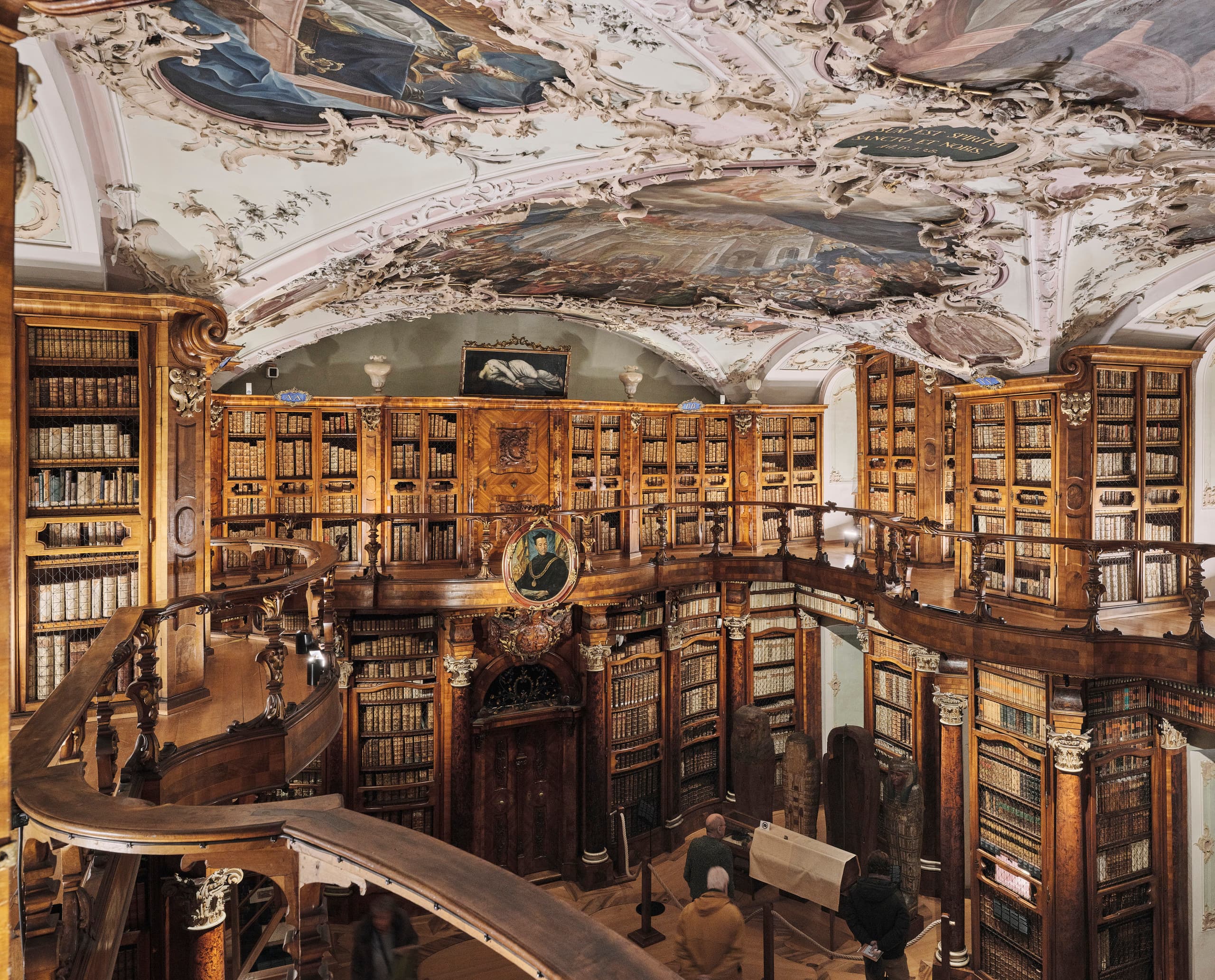 Prächtiger historischer Bibliothekssaal mit hohen Holzregalen voller Bücher und einer reich verzierten, gemalten Decke.