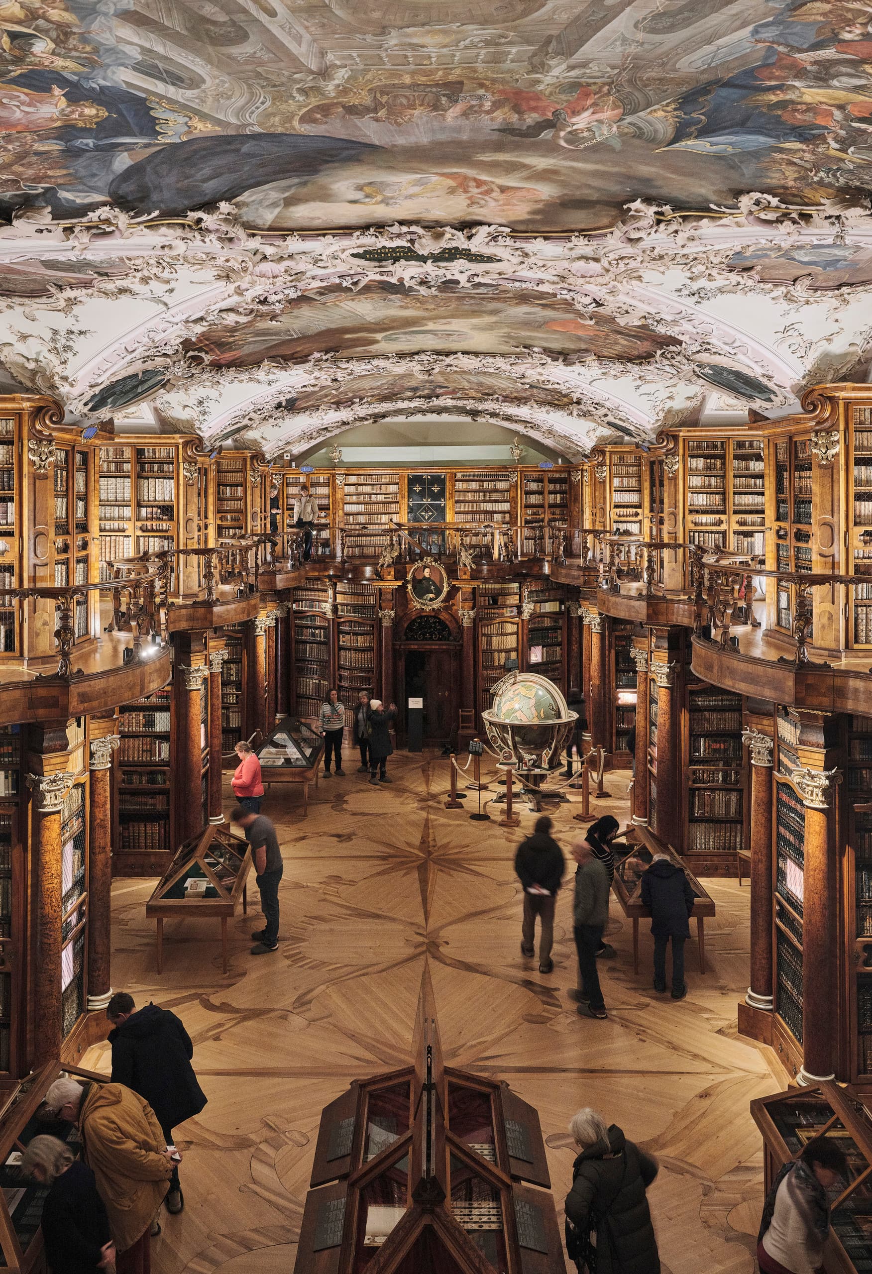 Historischer Bibliothekssaal mit hohen Holzregalen, kunstvoll bemalter Decke, großen Globen und Besucherinnen und Besuchern, die durch die Ausstellung gehen.