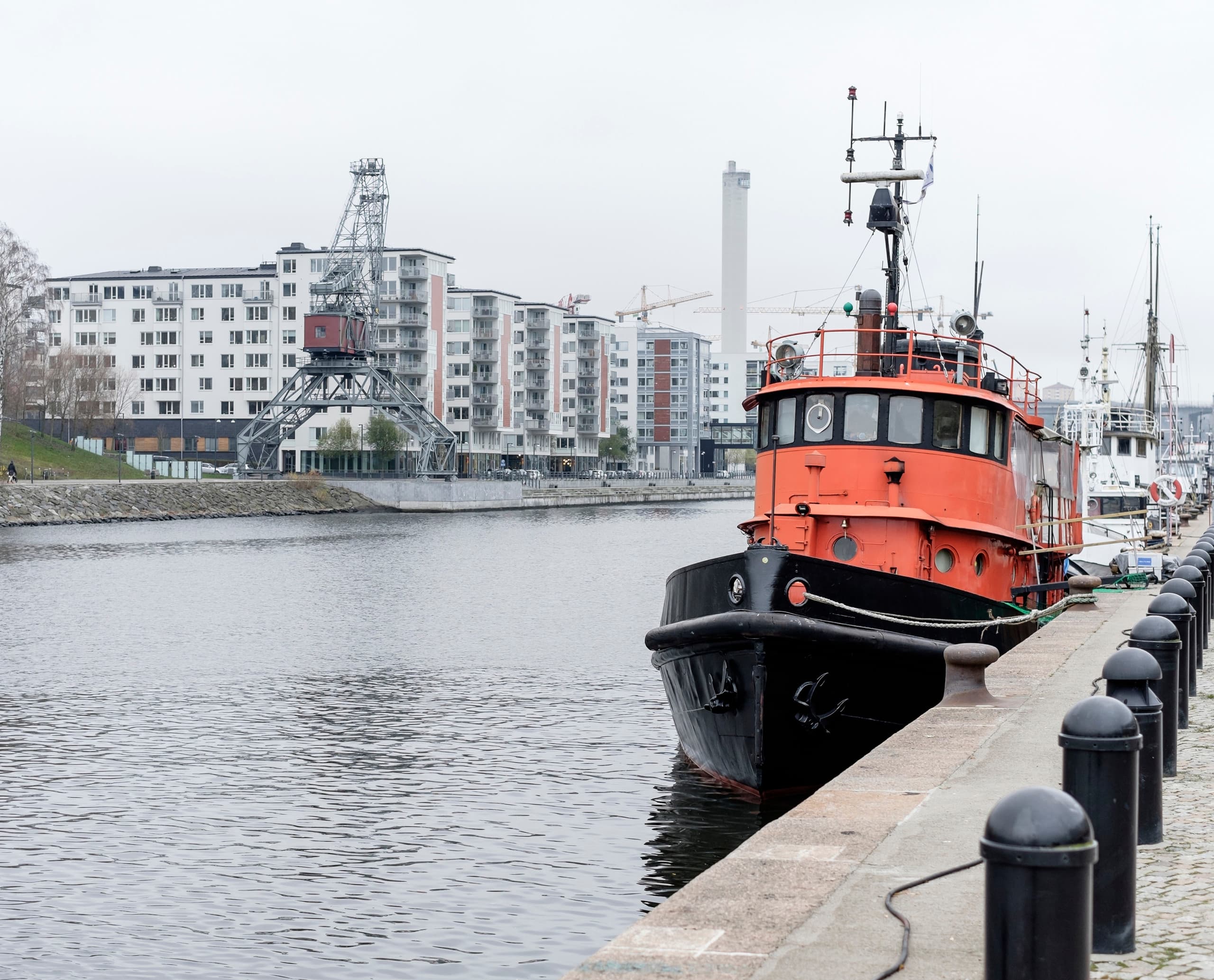 Ein rotes Lotsenboot liegt im Hafen von Stockholm, Schweden, vor dem Hintergrund von Wohngebäuden und einem Kran.
