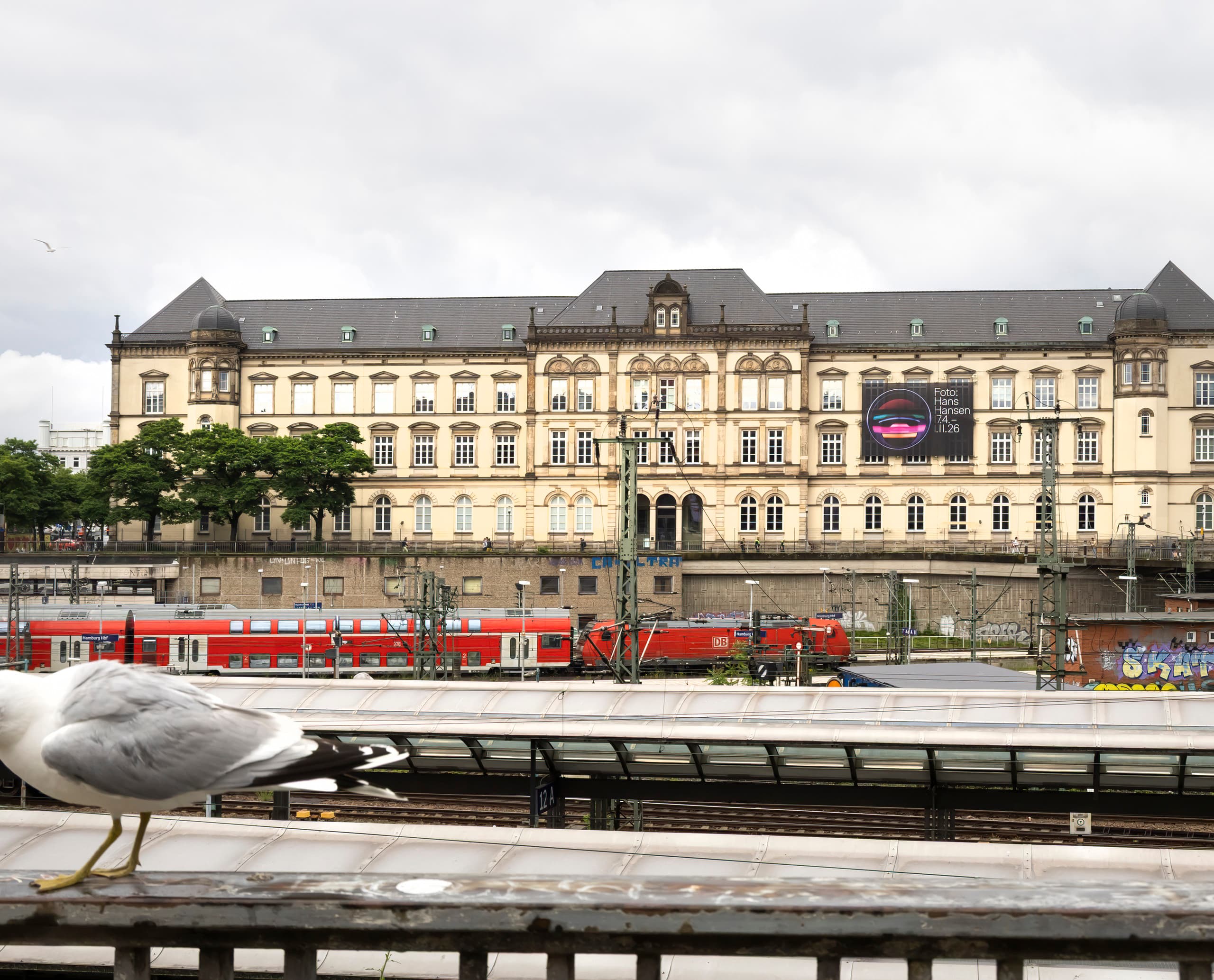 Außenansicht eines großen Gebäudes, Zugschienen im Vordergrund.