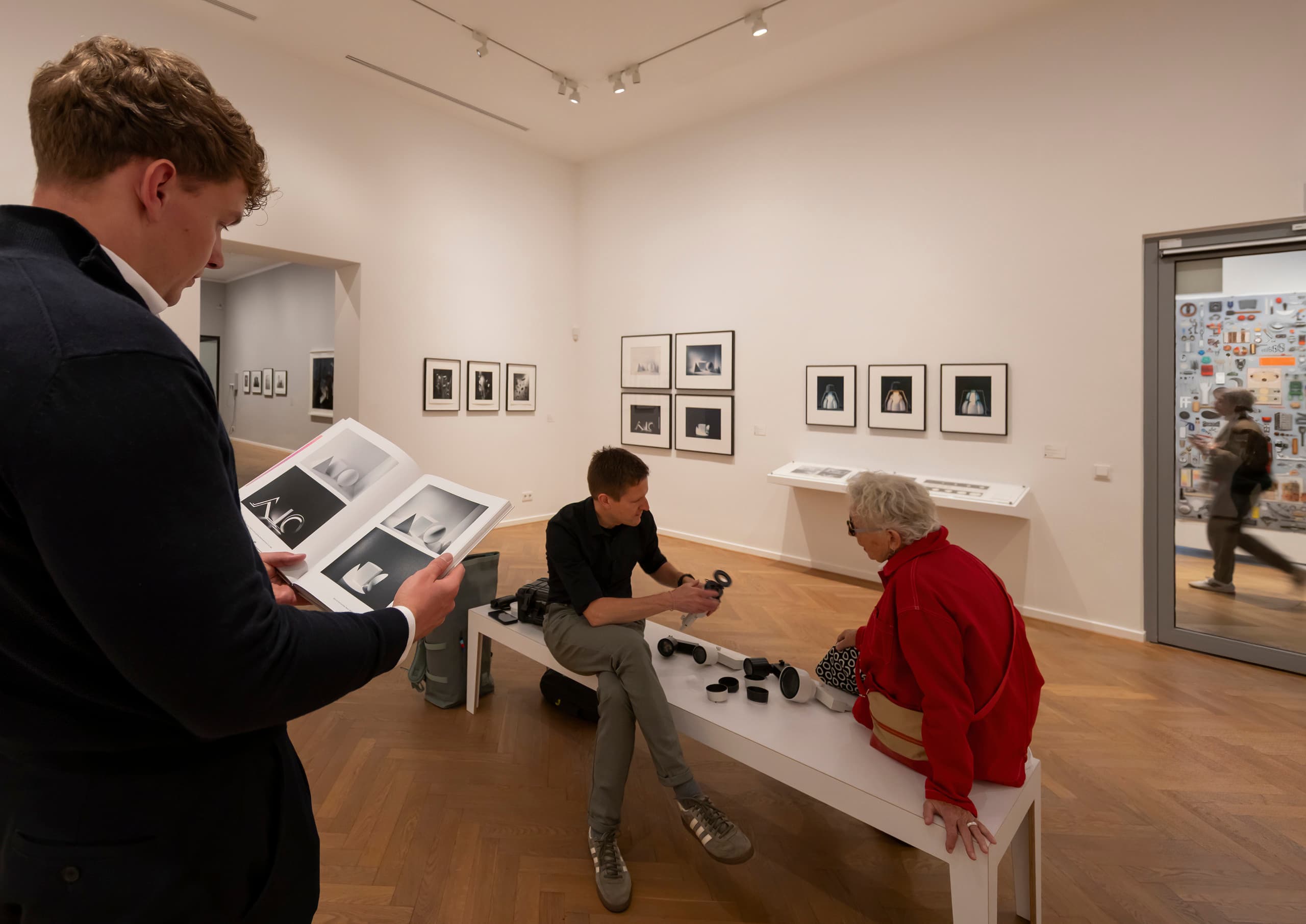 Ausstellungsraum, mittig auf einer Bank sitzen Menschen und begutachten Leuchtenkomponenten.