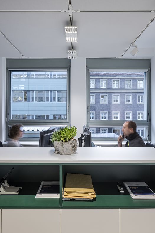 A detailed view of an office workstation positioned in front of a window front, featuring a continuous power rail and precisely aligned ceiling spotlights for zoned lighting at the desk.