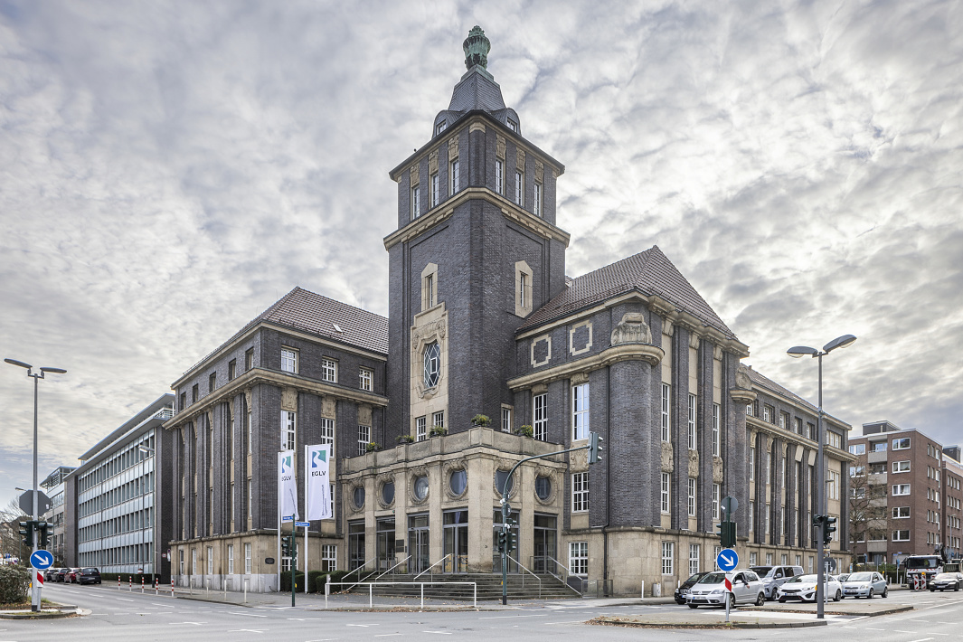 A historic brick administrative building with a striking tower on a street corner, photographed in an urban setting under a cloudy sky.