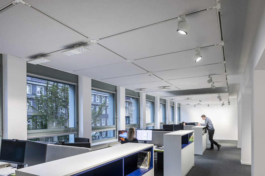 A modern open-plan office with floor-to-ceiling windows, spacious workstations and a continuous track lighting system with adjustable ceiling spotlights for flexible, zoned lighting.