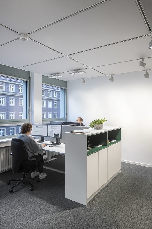 An open-plan office with two computer workstations by the windows and a continuous power rail with adjustable ceiling spotlights for precisely tailored workplace lighting.