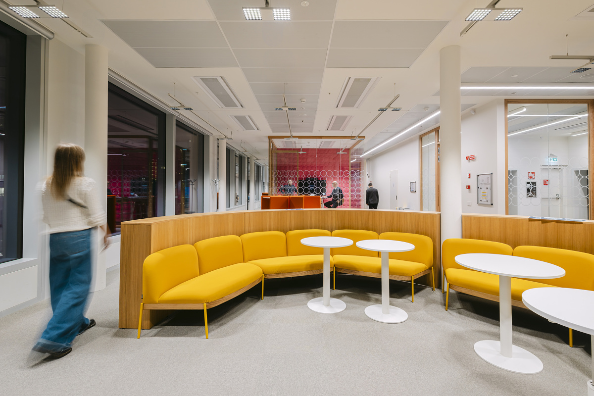 Modern ingerichte kantoorruimte met gebogen gele zitbank en witte, ronde tafels op de voorgrond; daarachter de beglaasde vergaderruimte met rode wandbekleding en personen die zich door de ruimte verplaatsen.