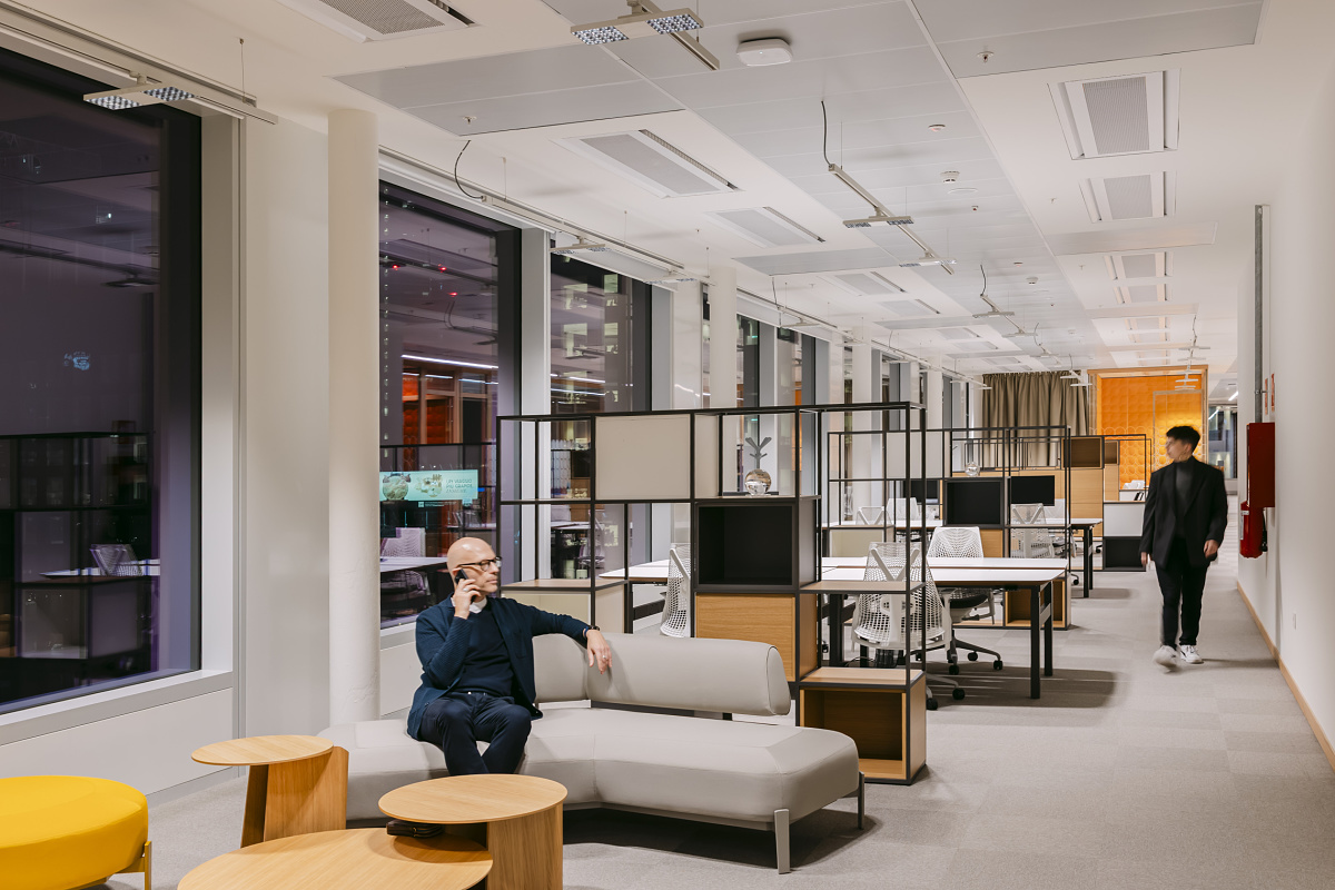 Moderne kantoorruimte met vensters die doorlopen tot op de vloer, lichte werkplekken en een loungebank op de voorgrond; op de achtergrond zijn meerdere bureaus met monitors te zien, terwijl een persoon zich door de ruimte verplaatst.