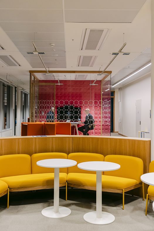 Modern vormgegeven kantoorruimte met gele zitbanken en witte, ronde tafels op de voorgrond. Daarachter de beglaasde vergaderruimte met rode wandbekleding en twee personen aan een tafel.