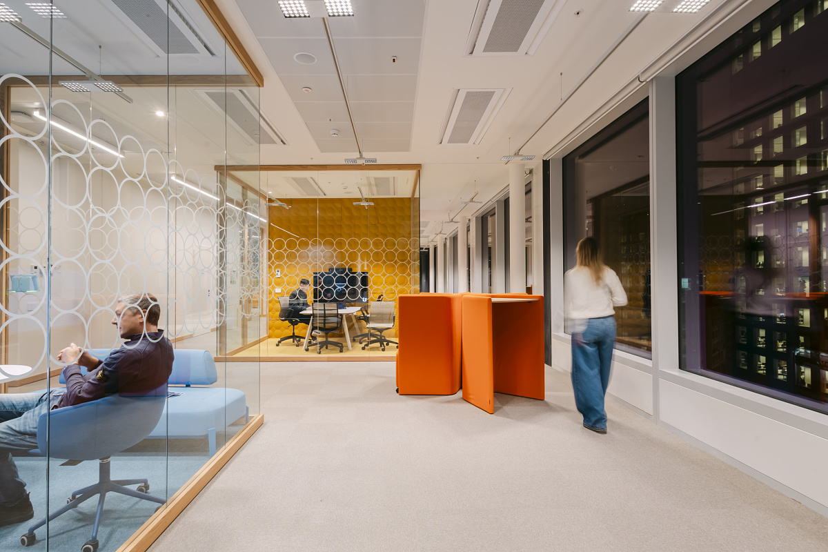 Bright office area with glass-walled meeting rooms, decorative circular-pattern‑ glazing and contemporary seating zones. In the foreground, one person sits in a blue armchair; in the background, another meeting room with a yellow wall is visible while a person walks past the window.
