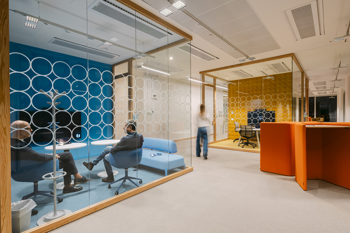 Modern office area with two glass-walled meeting rooms, one blue and one yellow, both featuring circular-pattern glazing. In the front room, two people sit in blue armchairs; in the rear room, there is a large table, while another person walks along the corridor.