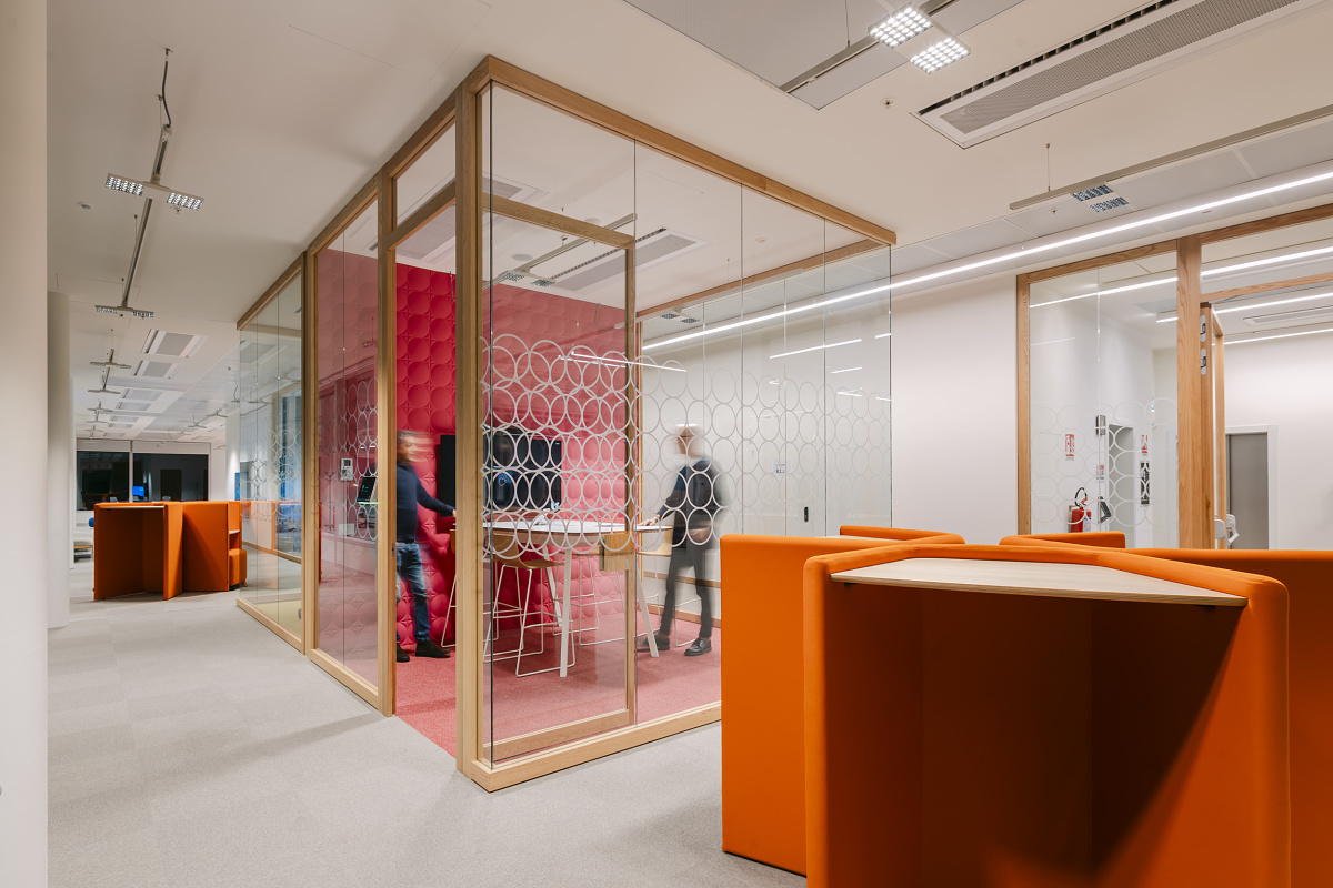 Modern office area with a glass-walled meeting room featuring circular-pattern glazing. Inside, people stand around a high table, while orange standing workstations are visible ‑in the foreground.