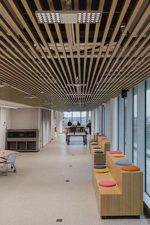 Large modern office space with a slatted timber ceiling, full-height windows and colored seating steps. In the background are a table tennis table and high stools, where two people are seated by the window.
