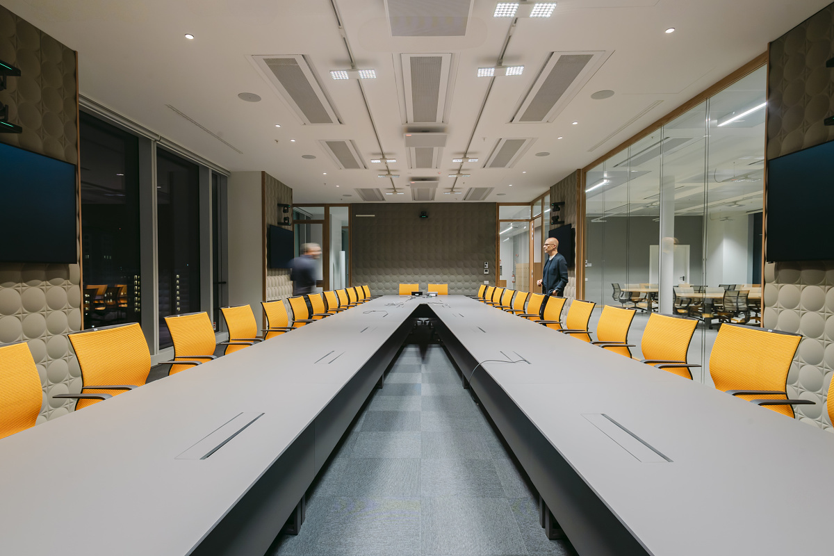 Large modern conference room with a U‑shaped table and numerous yellow swivel chairs. Large screens are positioned on the sides, while two people move through the room.