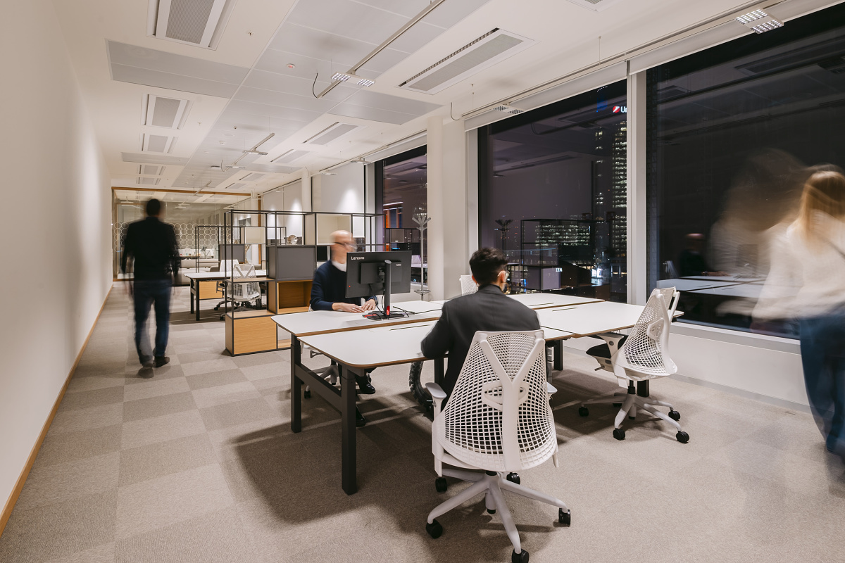 Bright modern office area with several desks and ergonomic office chairs. One person is seated at a workstation in front of a screen, while two others walk through the room. Large windows provide a view of the city at night.