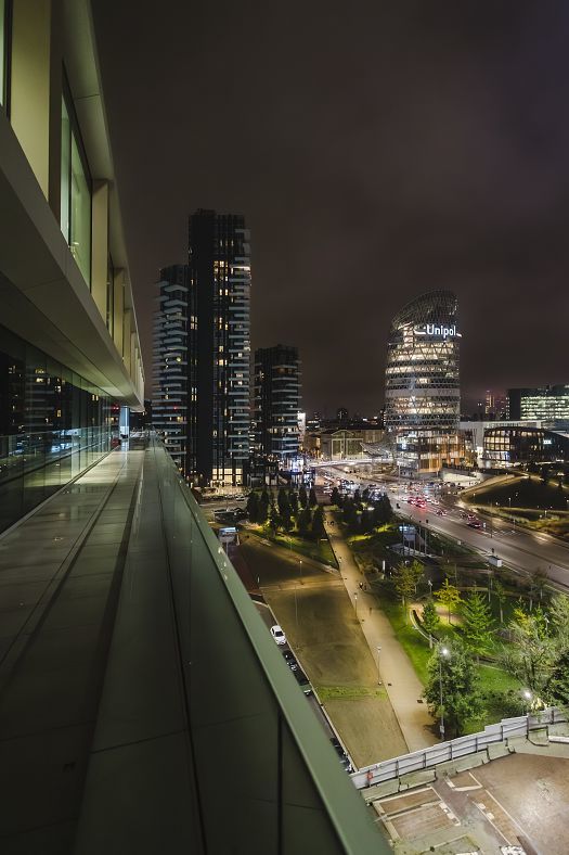 Night-time view from an elevated terrace with a glass balustrade, overlooking modern illuminated high-rise buildings and a lively cityscape with streets and green spaces.