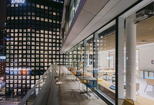 Night-time view of a modern office terrace with a glass balustrade and colored chairs along the glazed facade. On the right, a bright interior seating area is visible; on the left stands a brightly lit office tower with numerous windows.