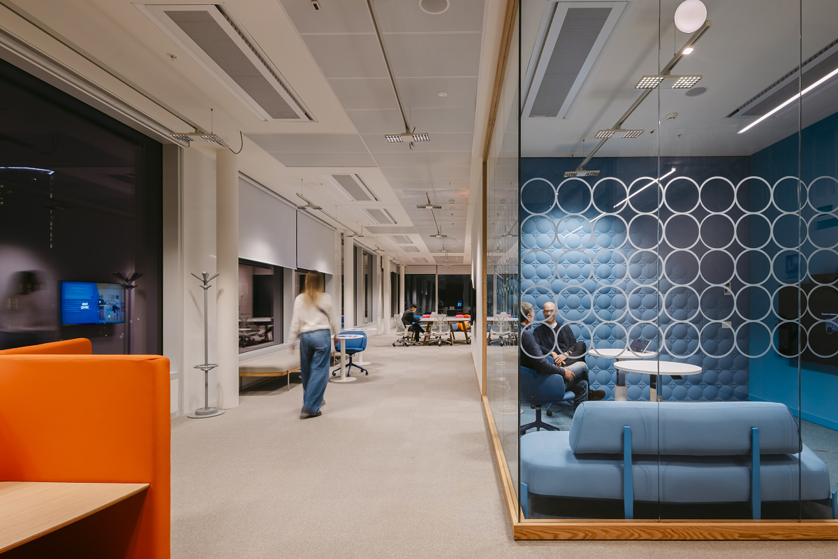 Modern office interior with an open-plan workspace and a glass-walled meeting room featuring a blue upholstered wall. Several people are seated or moving through the brightly lit room.